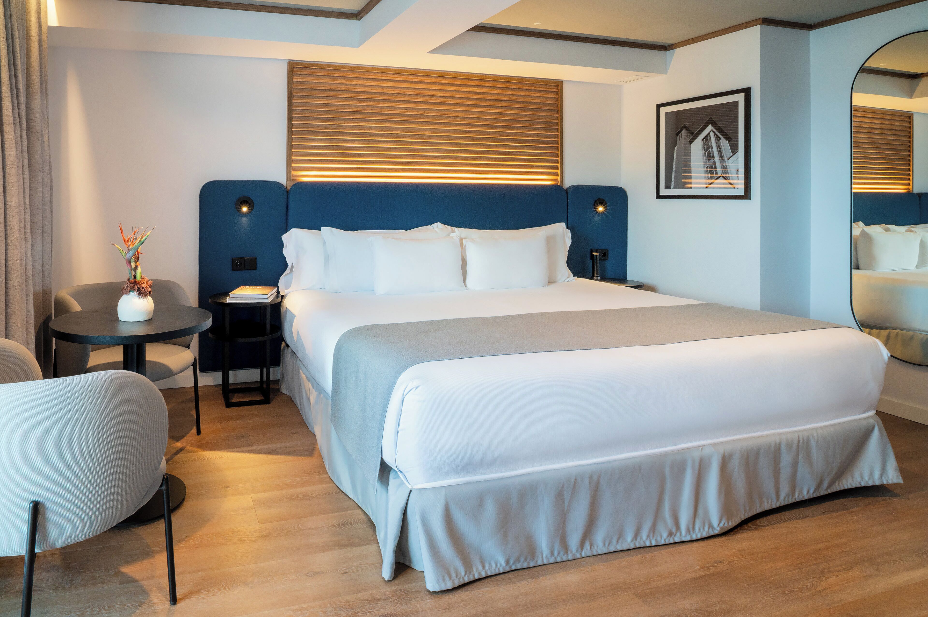 A hotel room with a large bed, a round table, a chair, a nightstand, and a mirror.