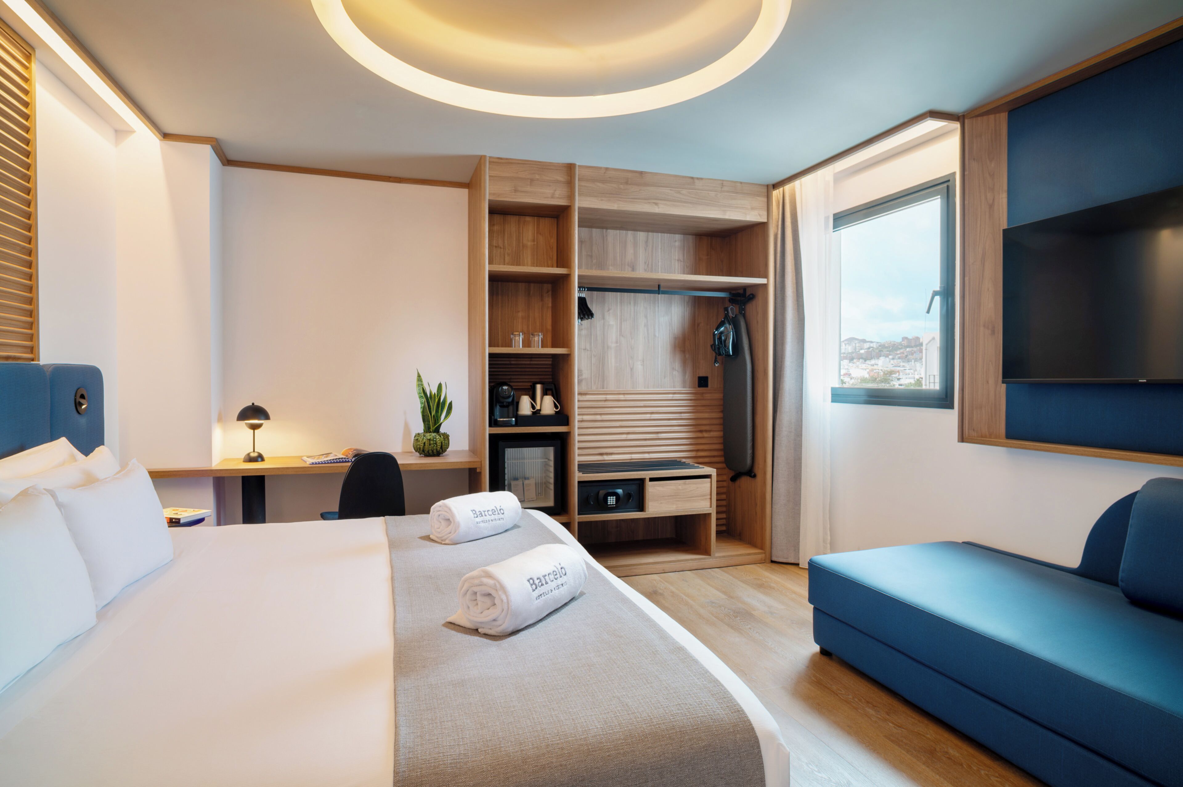 A modern hotel room with a bed, a desk, a wardrobe, a sofa, and a TV.