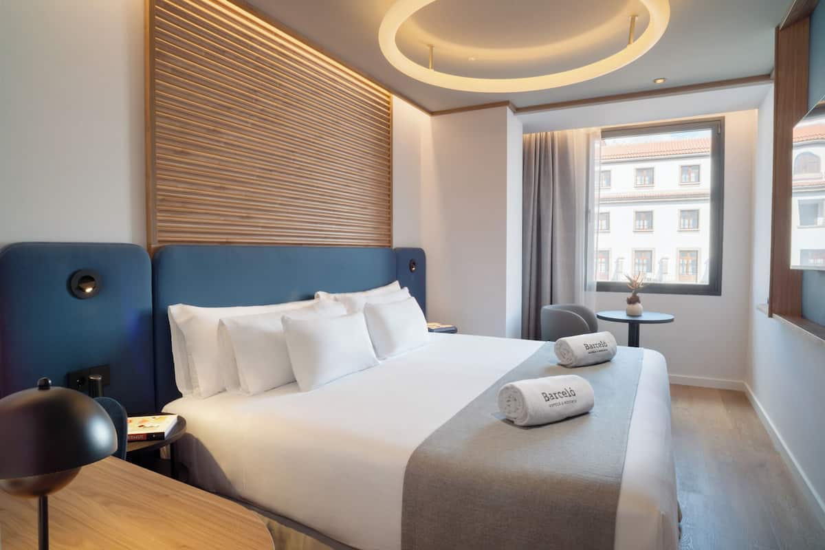 Habitación de hotel con una cama grande, una mesita, una silla, una lámpara y una ventana con vistas a edificios.