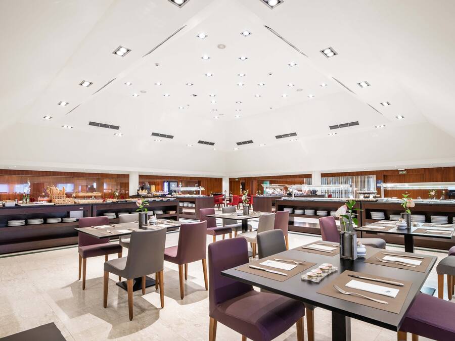 A spacious restaurant with multiple dining tables and chairs, a kitchen area in the background, and a high ceiling with recessed lighting.