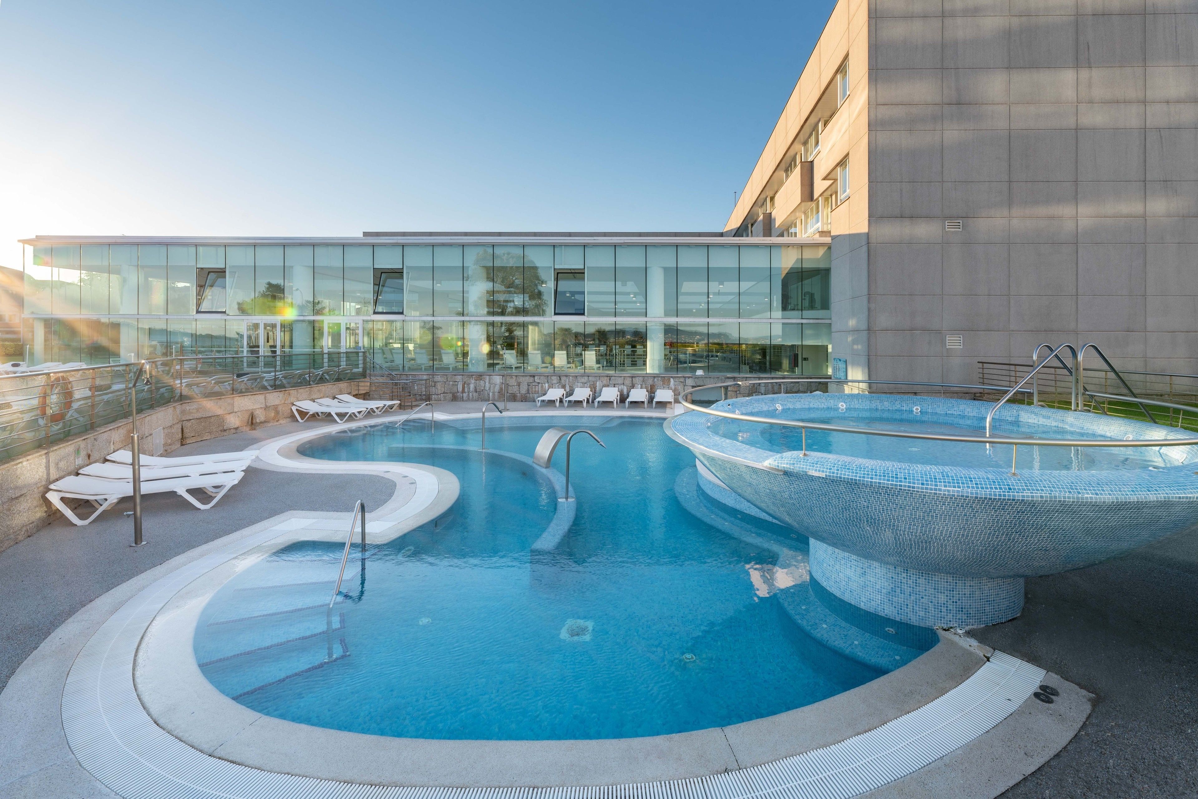 Una piscina con cerramiento de vidrio, sillas de descanso y un edificio moderno al fondo.