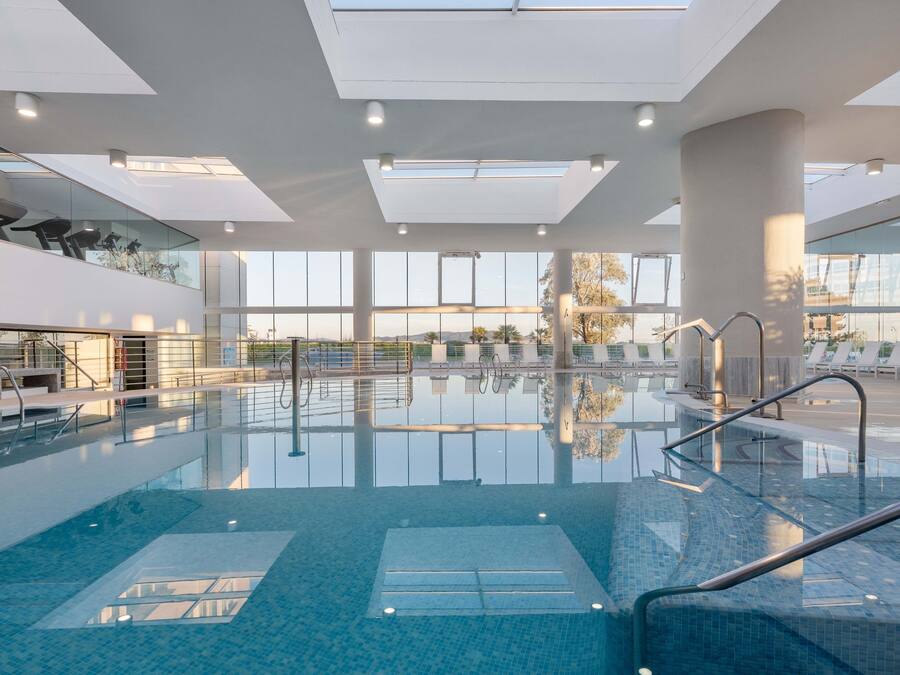 A modern indoor swimming pool with clear glass walls, a sunlit ceiling, and a view of the outdoors.
