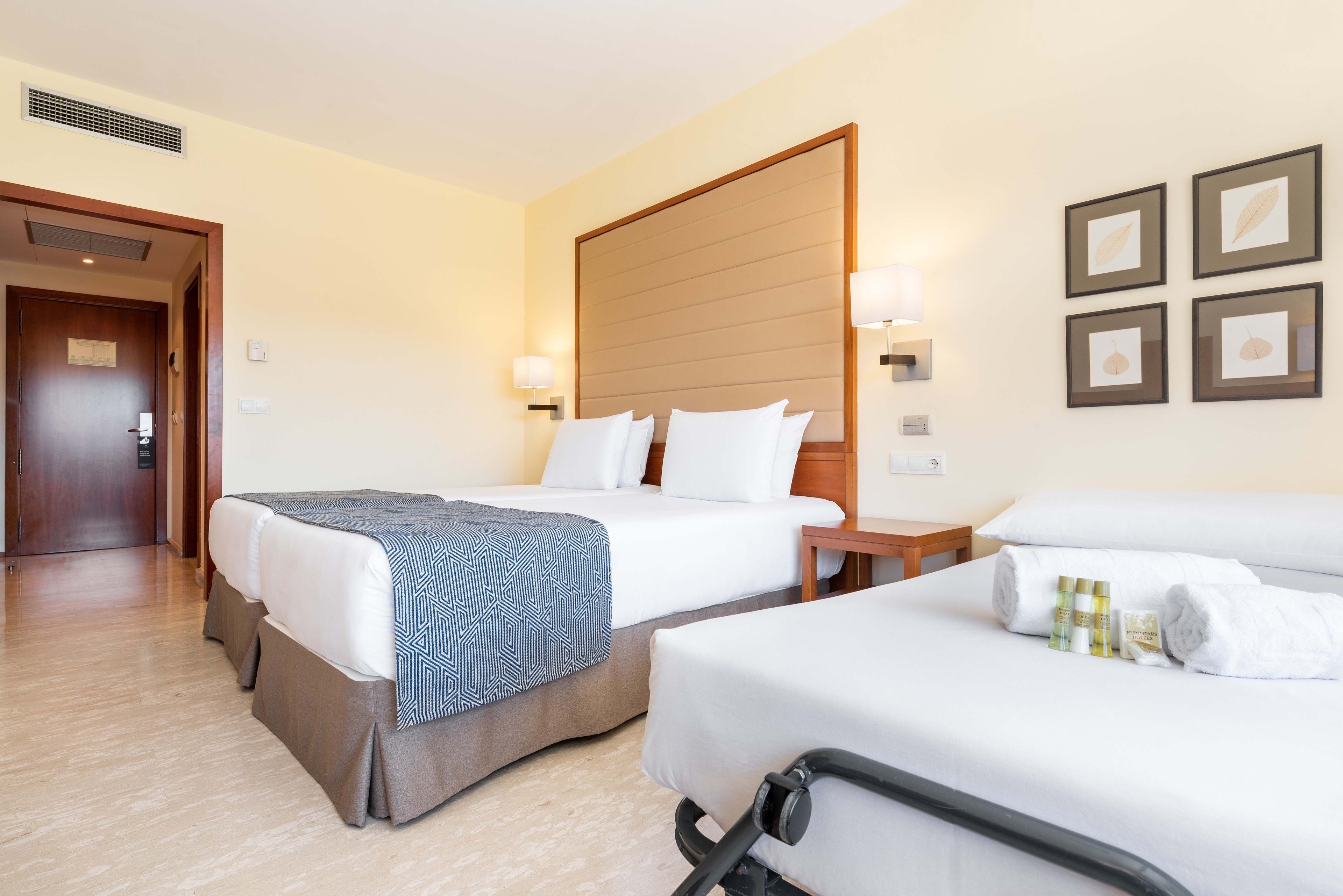 Habitación de hotel con dos camas, cabecero de madera, lámparas de noche y cuadros en la pared.