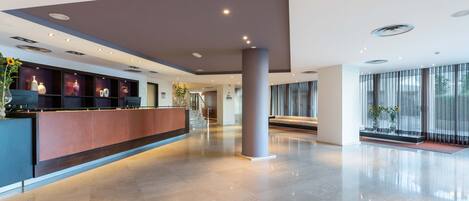 A modern hotel lobby with a reception desk, a staircase, and large windows.