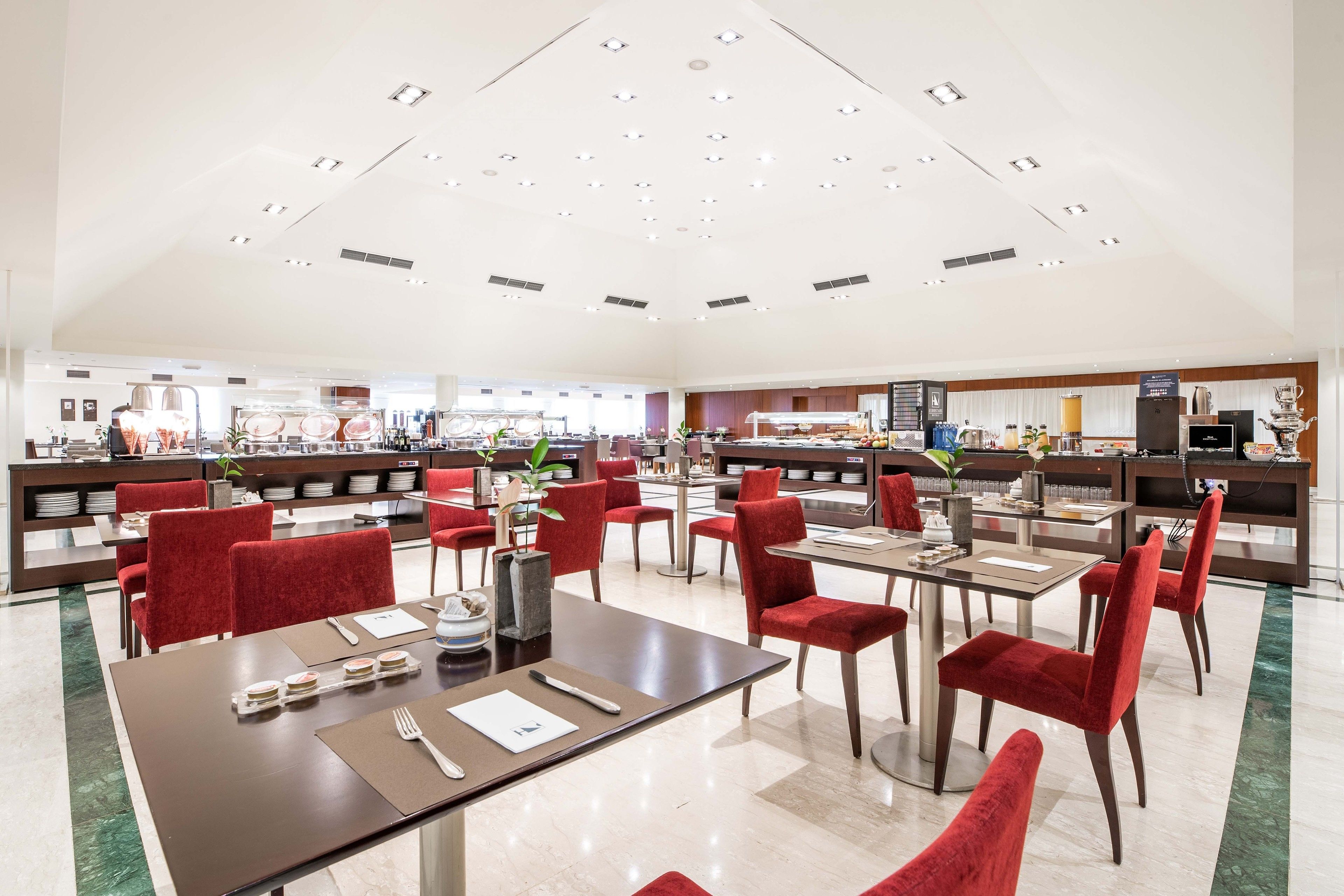 A spacious dining area with red upholstered chairs and tables, a buffet counter in the background, and a modern ceiling design.