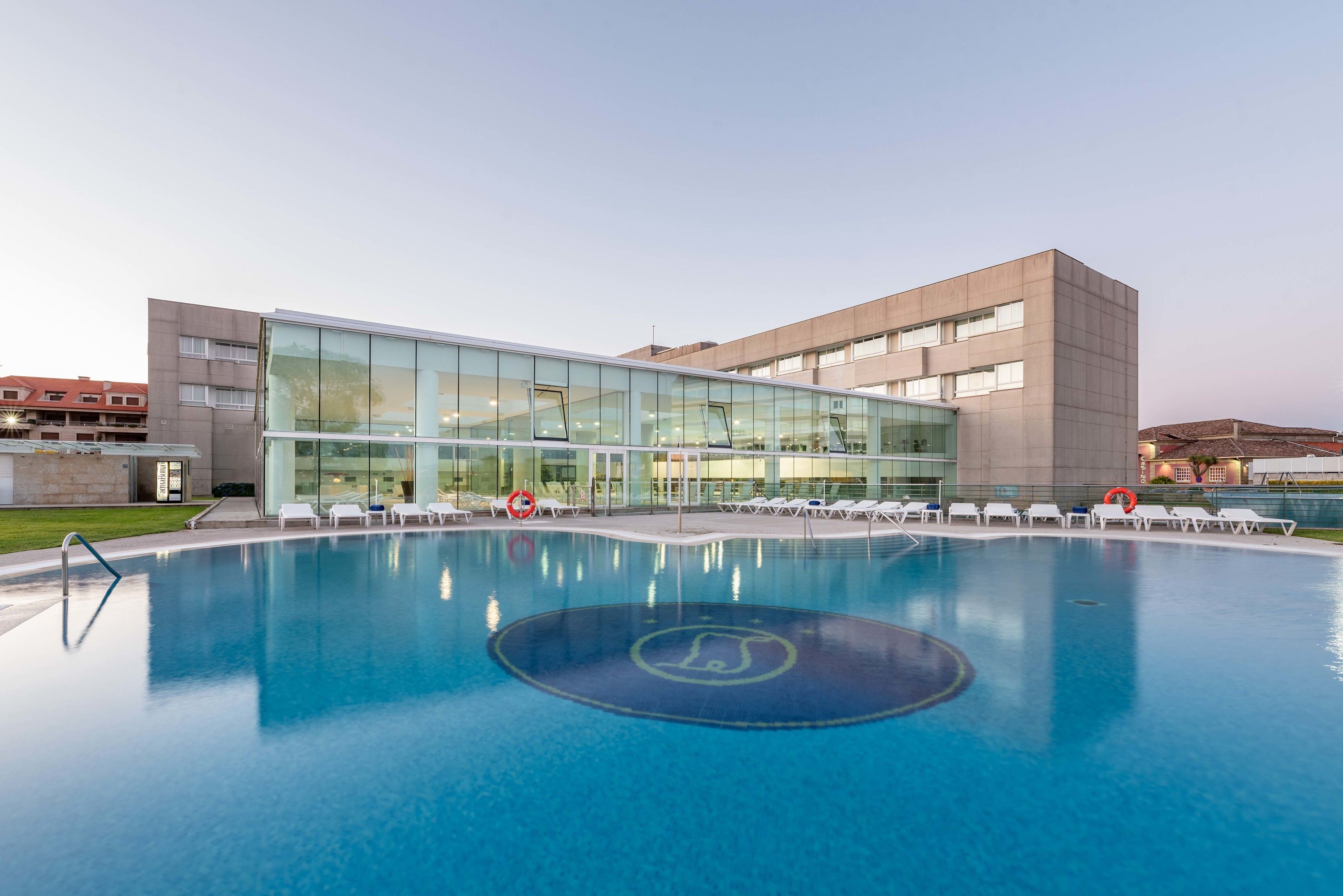 Un bâtiment moderne avec une piscine à l’avant, entouré de chaises longues.