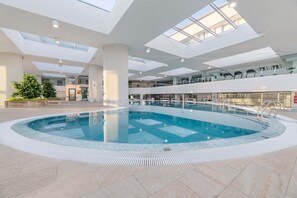 Een binnenzwembad met een helder uitzicht op de lucht door middel van dakramen, omgeven door een ruim atrium met meerdere niveaus en zitgedeeltes.