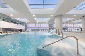 Een modern binnenzwembad met dakramen, lounge stoelen en uitzicht op de stad.