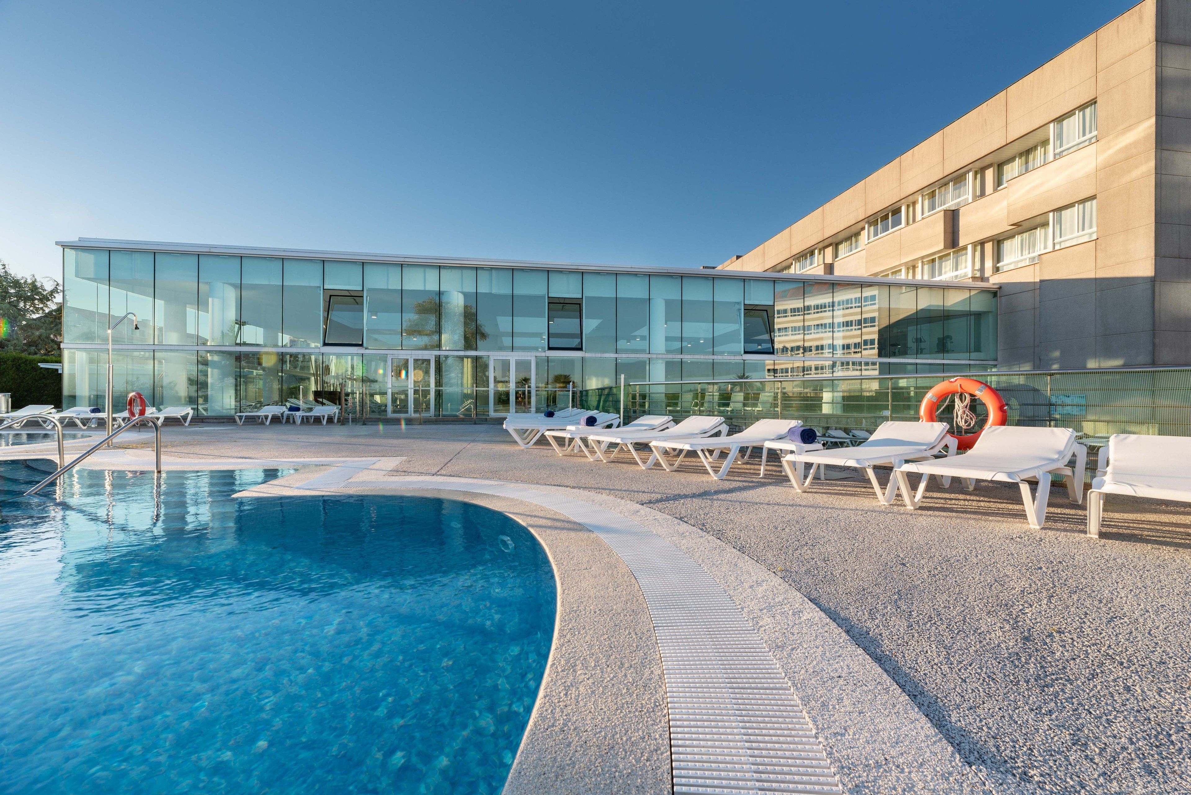 Un hôtel moderne doté d’une façade en verre, d’une piscine et de chaises longues.