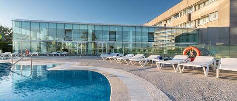 Un hotel moderno con fachada de vidrio, piscina y tumbonas.