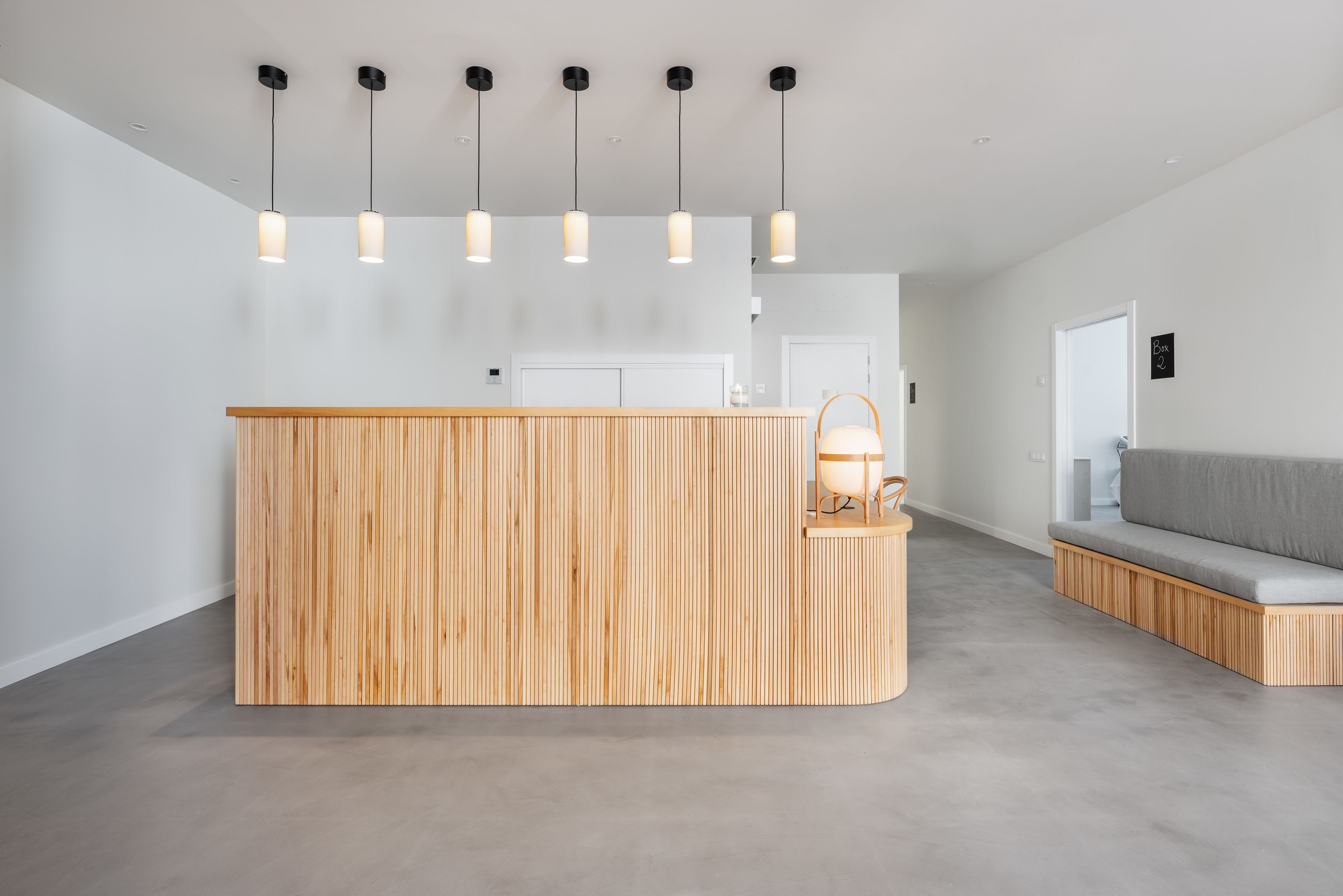 Un área de recepción con un mostrador de madera, iluminación colgante, un sofá gris y una puerta con un letrero.