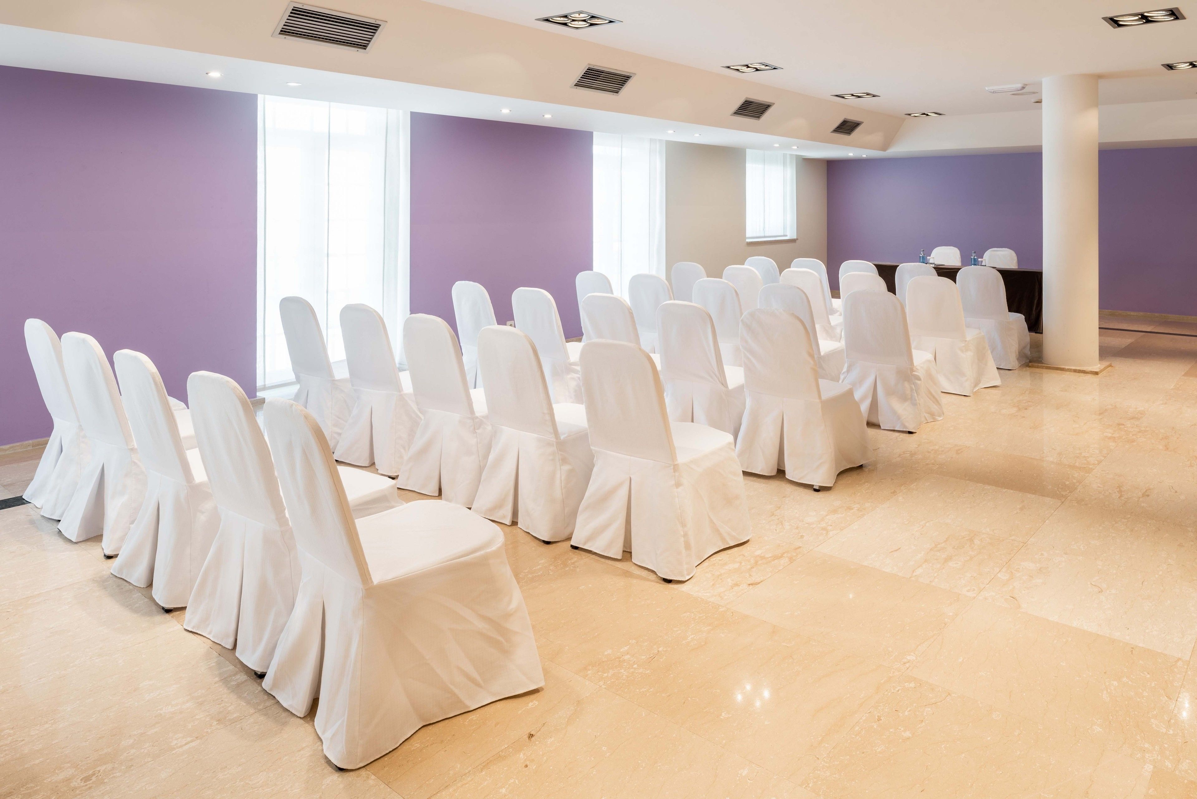 Un salón de conferencias con filas de sillas blancas y una mesa negra, ventanas amplias y una pared morada.