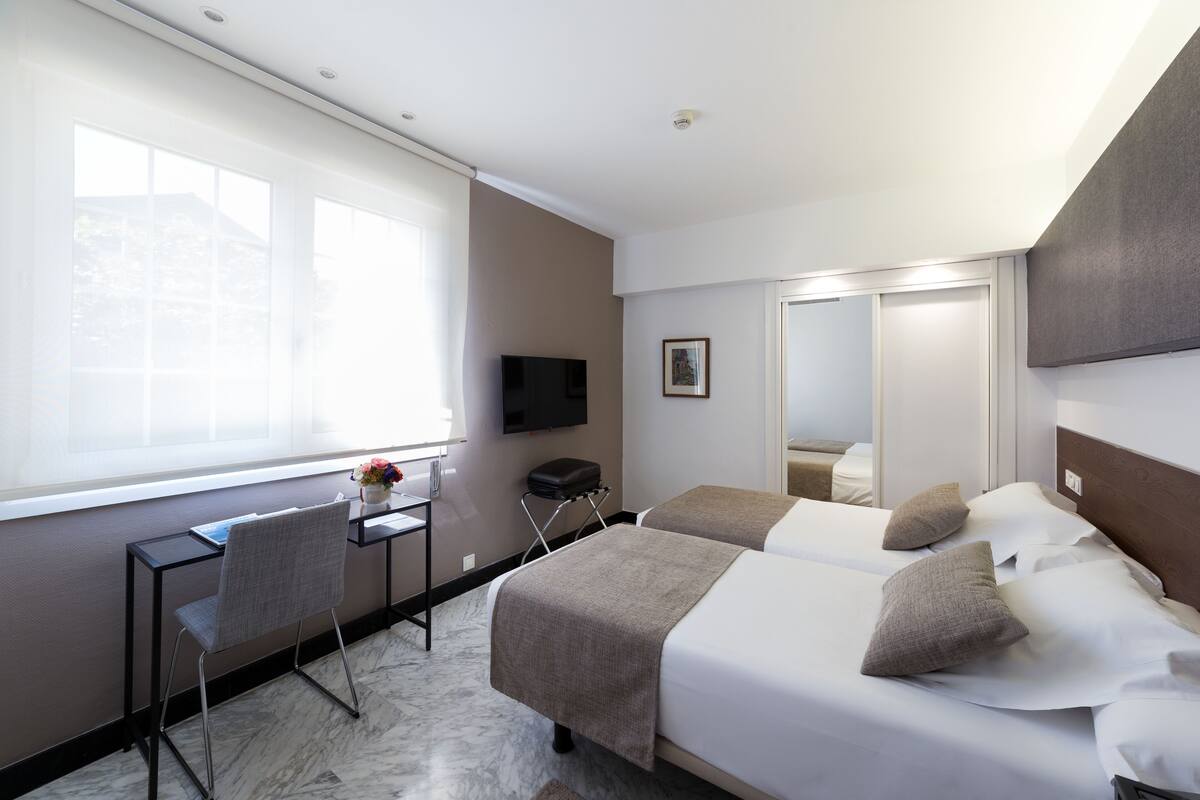 A hotel room with two beds, a desk, a chair, a TV, and a large window.