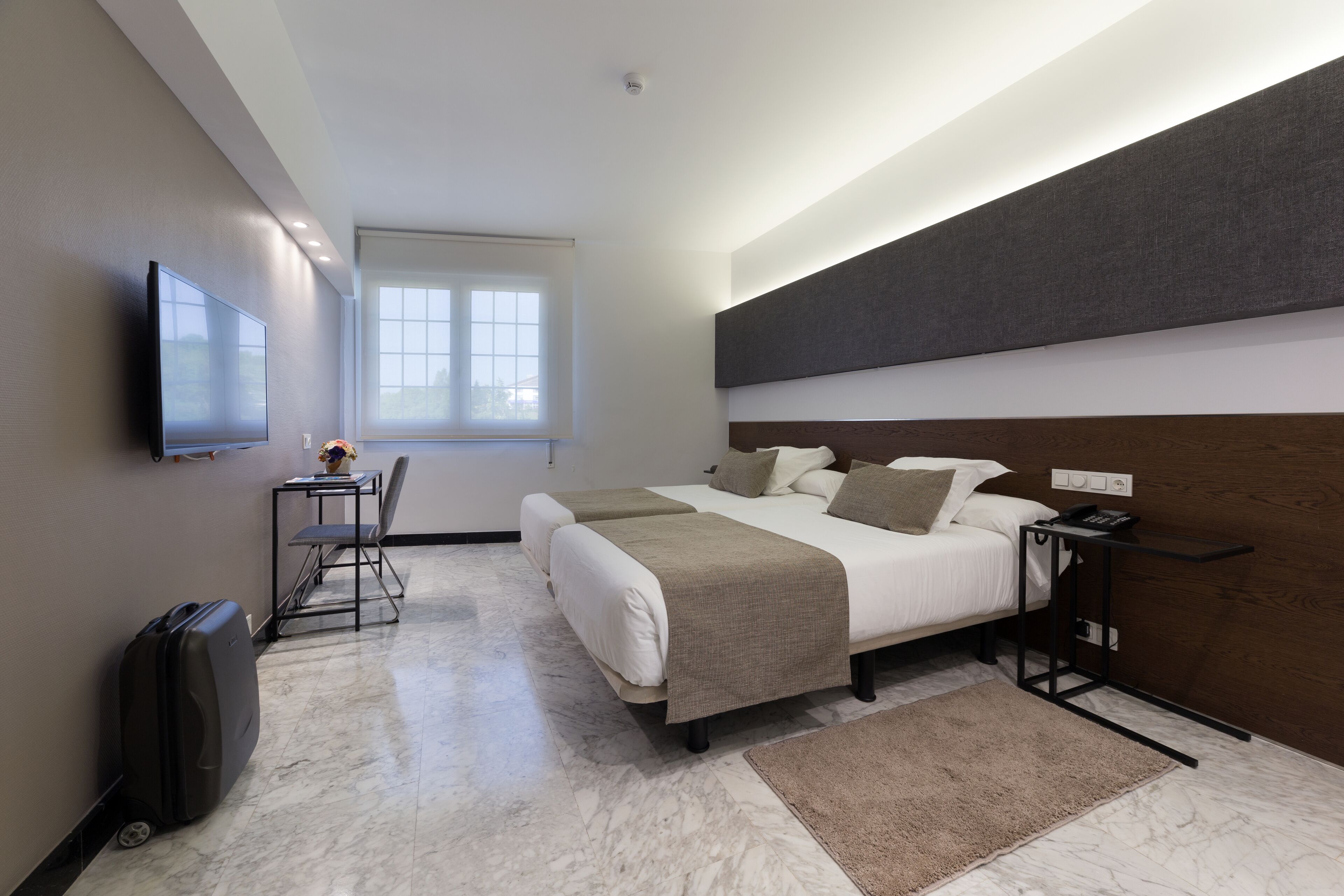 A modern hotel room with two beds, a flat-screen TV, a small table with a chair, a desk with a lamp, and a suitcase.