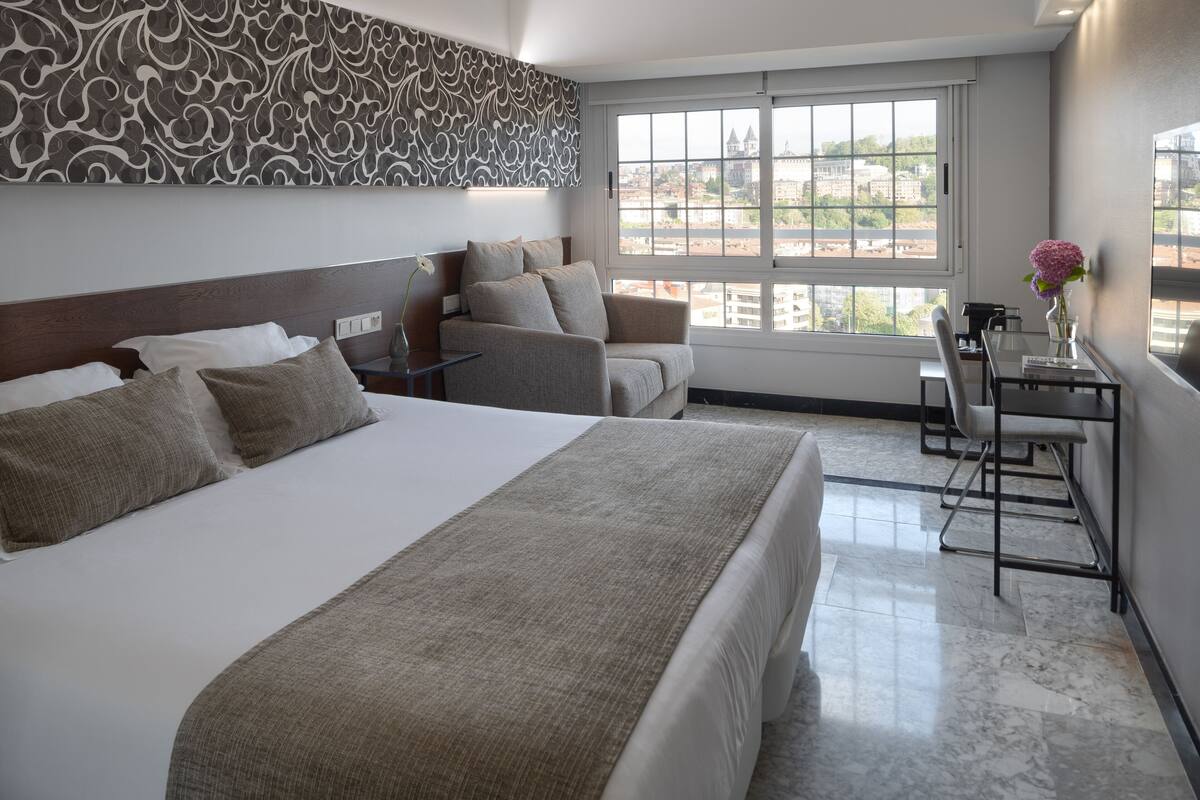 A hotel room with a large bed, a sofa, a desk with a chair, and a window with a city view.