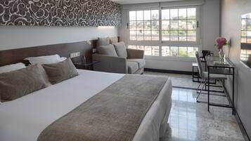 A hotel room with a large bed, a sofa, a desk with a chair, and a window with a city view.