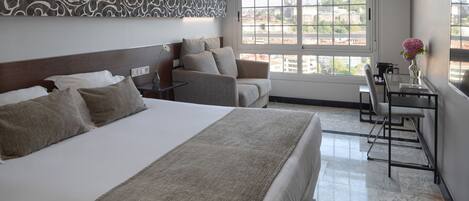 A hotel room with a large bed, a sofa, a desk with a chair, and a window with a city view.