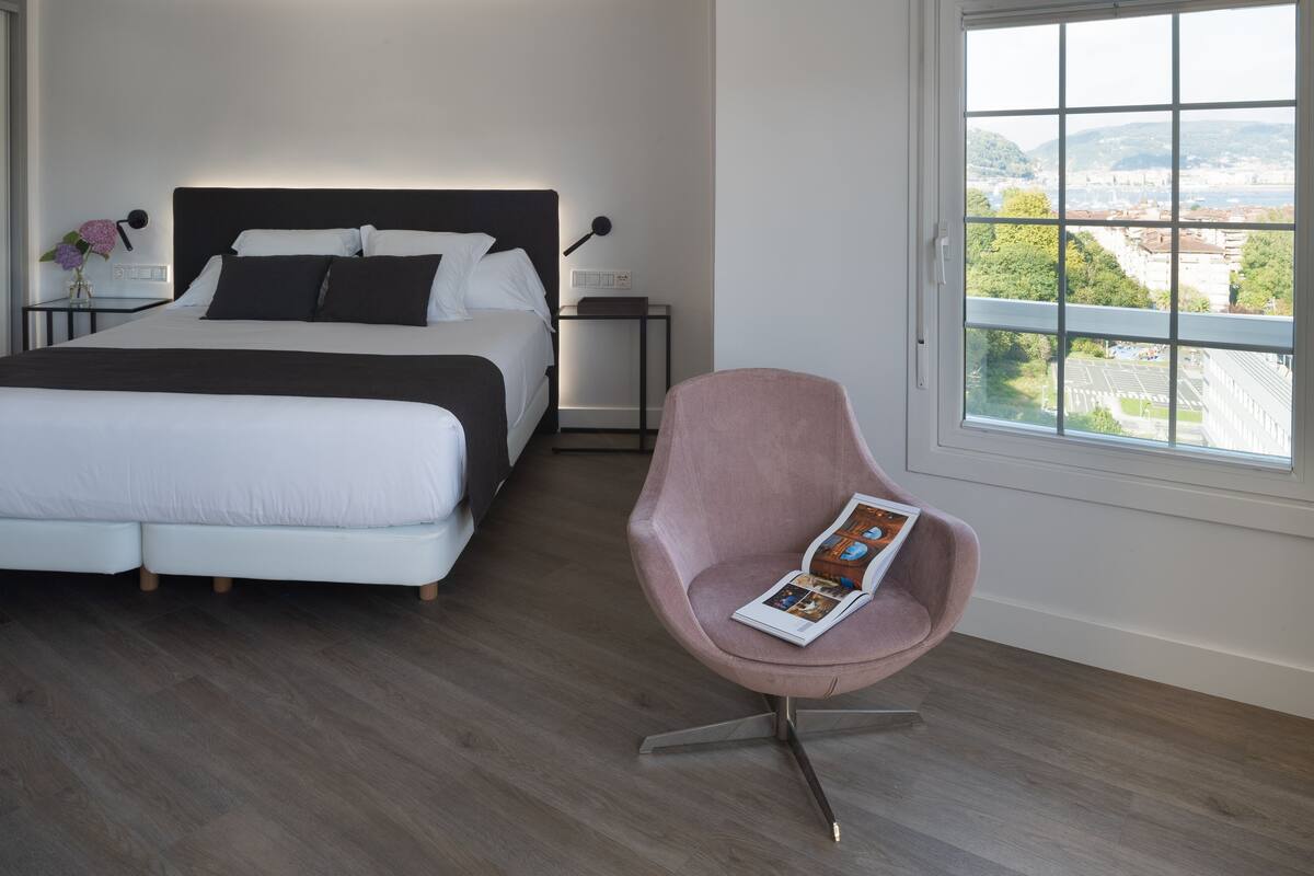 A bedroom with a bed, bedside tables, a chair, and a window with a city view.