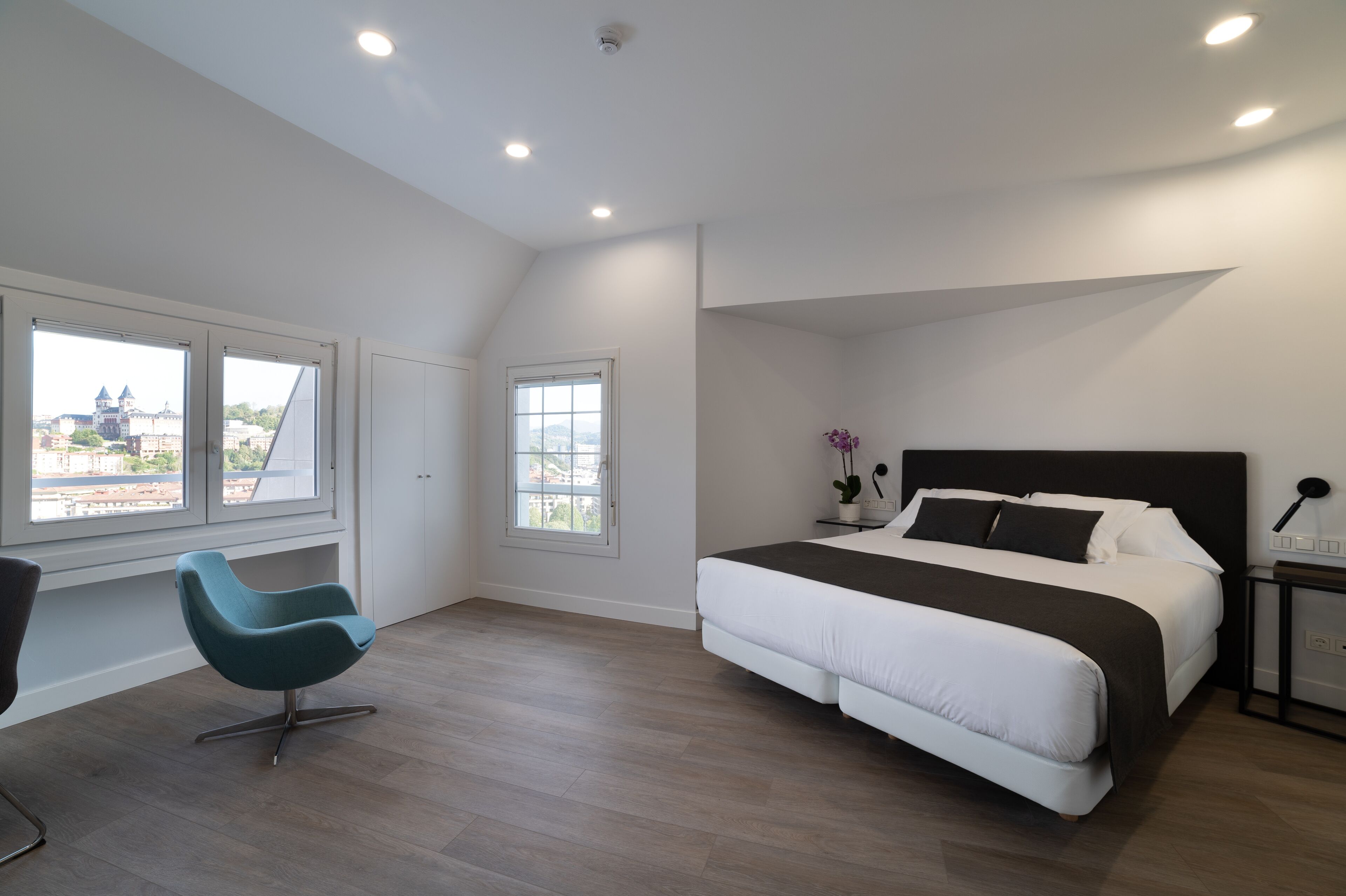 Un dormitorio moderno con una cama grande, una silla y dos ventanas con vistas a la ciudad.