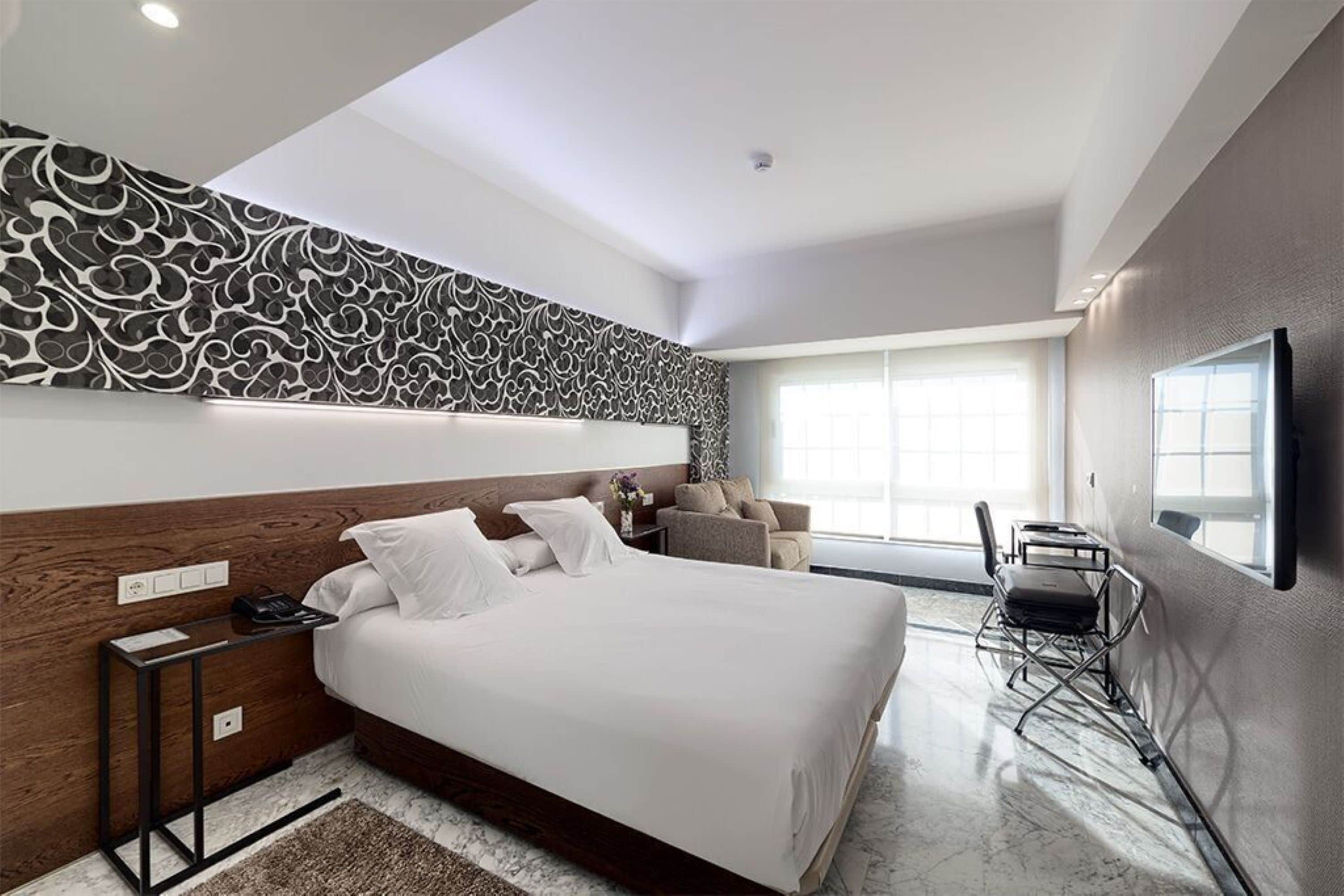 A modern hotel room with a large bed, a wooden headboard, a wall-mounted TV, and a seating area with a chair and table.