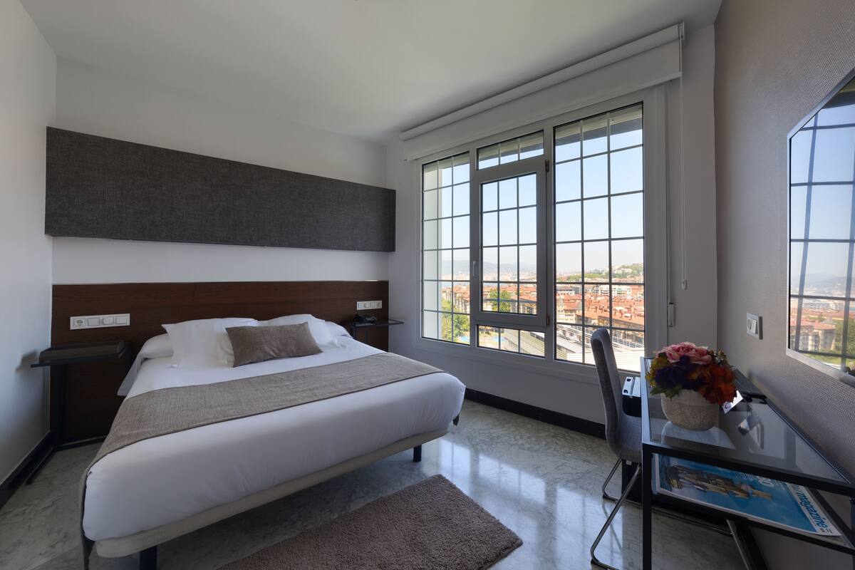 A modern hotel room with a large bed, a desk with a vase of flowers, and a view of the city through large windows.