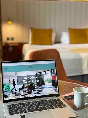 A laptop on a table with a hotel room in the background. The room has a bed with yellow pillows, a nightstand, and a lamp. The laptop screen displays a webpage with the text "An Address like no other".
