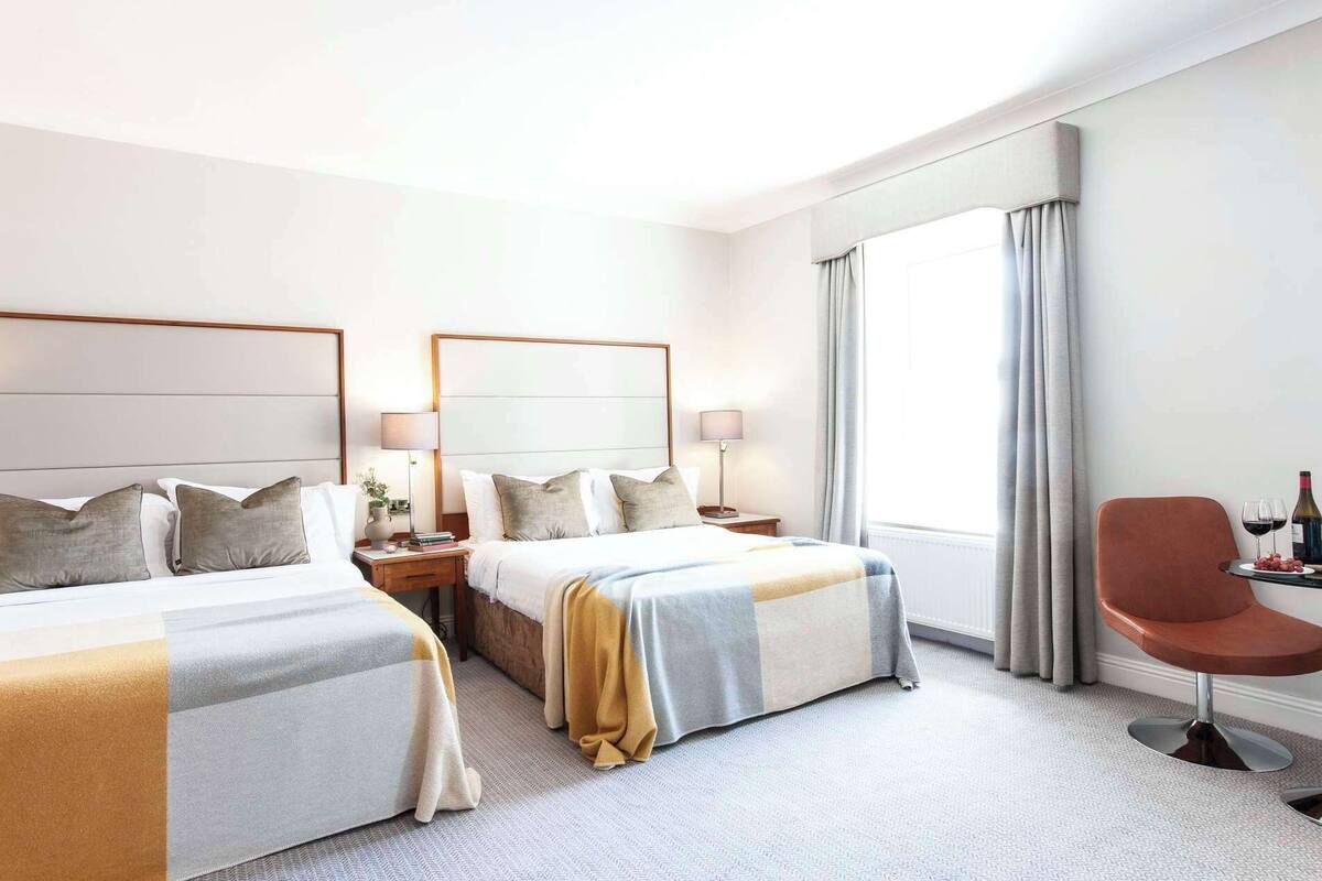 A hotel room with two beds, a sitting area with a chair, a table with a bottle and glasses, and a large window with curtains.