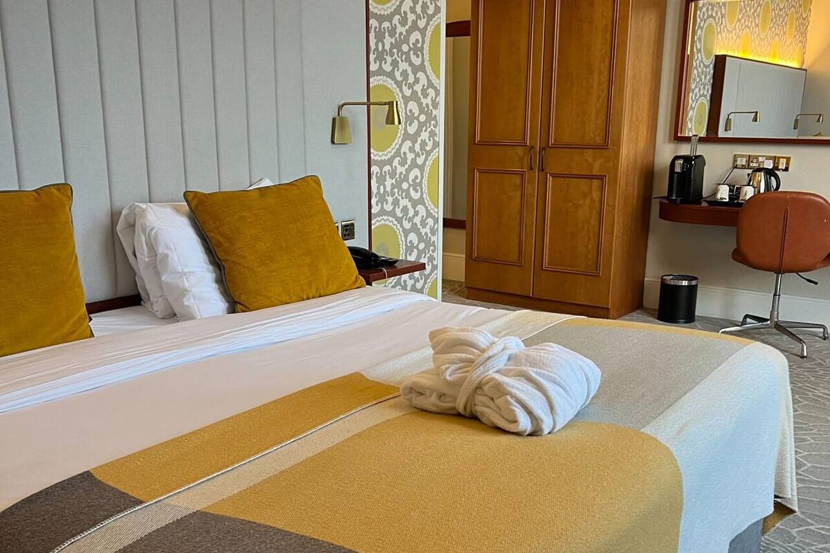 A hotel room with a large bed, two pillows, a wooden wardrobe, a desk with a chair, and a mirror.