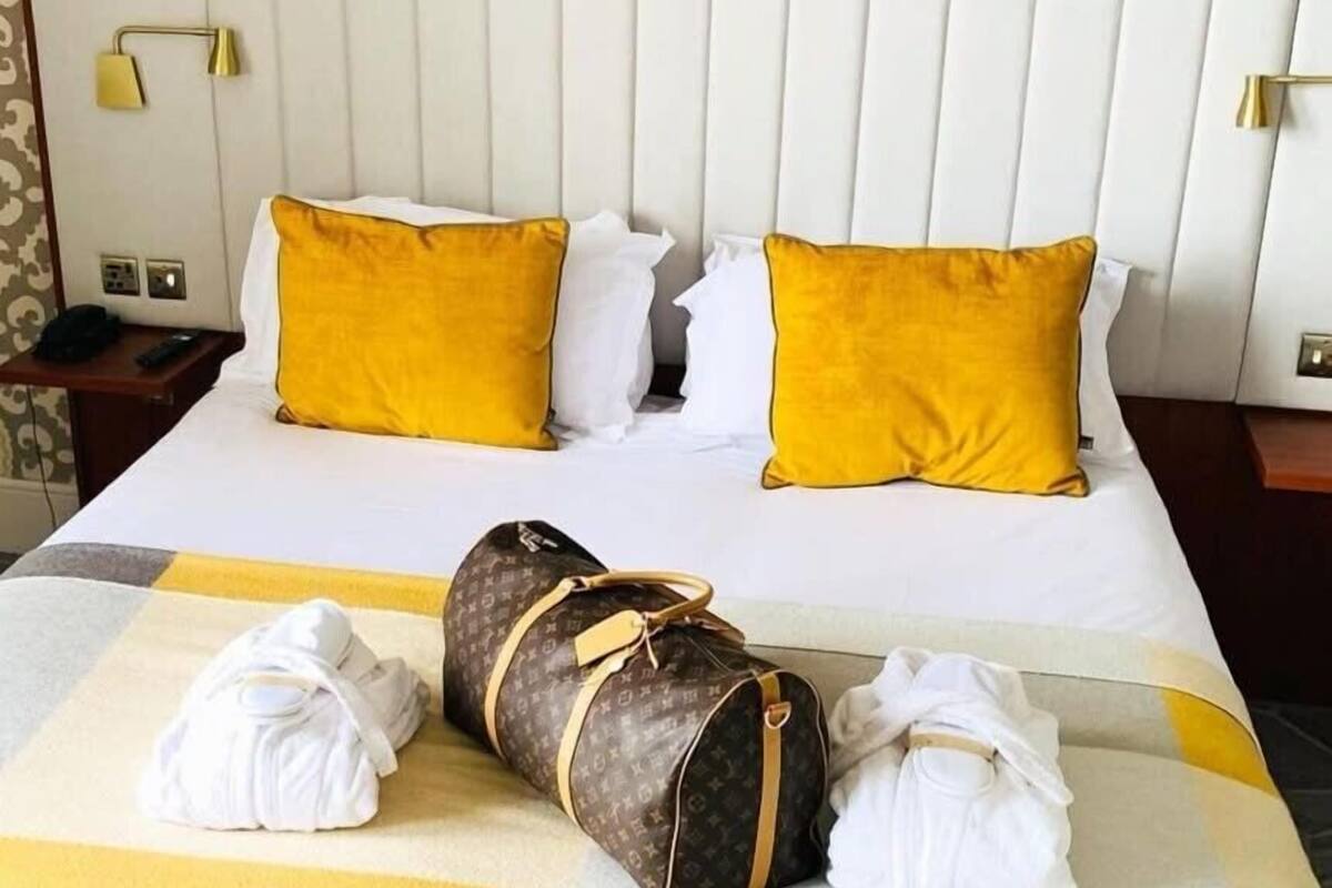 A hotel room with a bed, two yellow pillows, a white blanket, and a Louis Vuitton duffel bag.