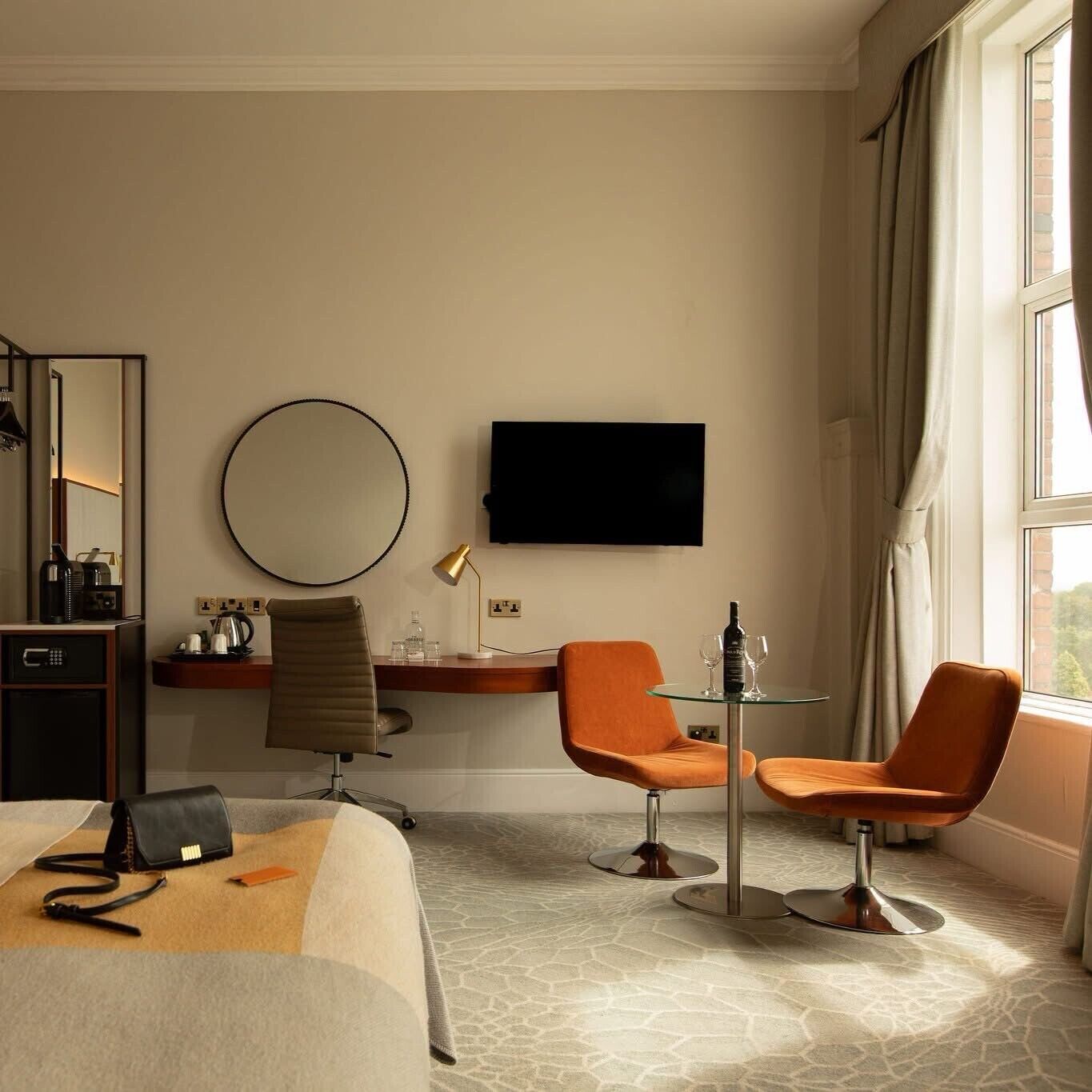 Une chambre d’hôtel comprenant un lit, un bureau, deux chaises, une télévision, un miroir et une fenêtre avec des rideaux.