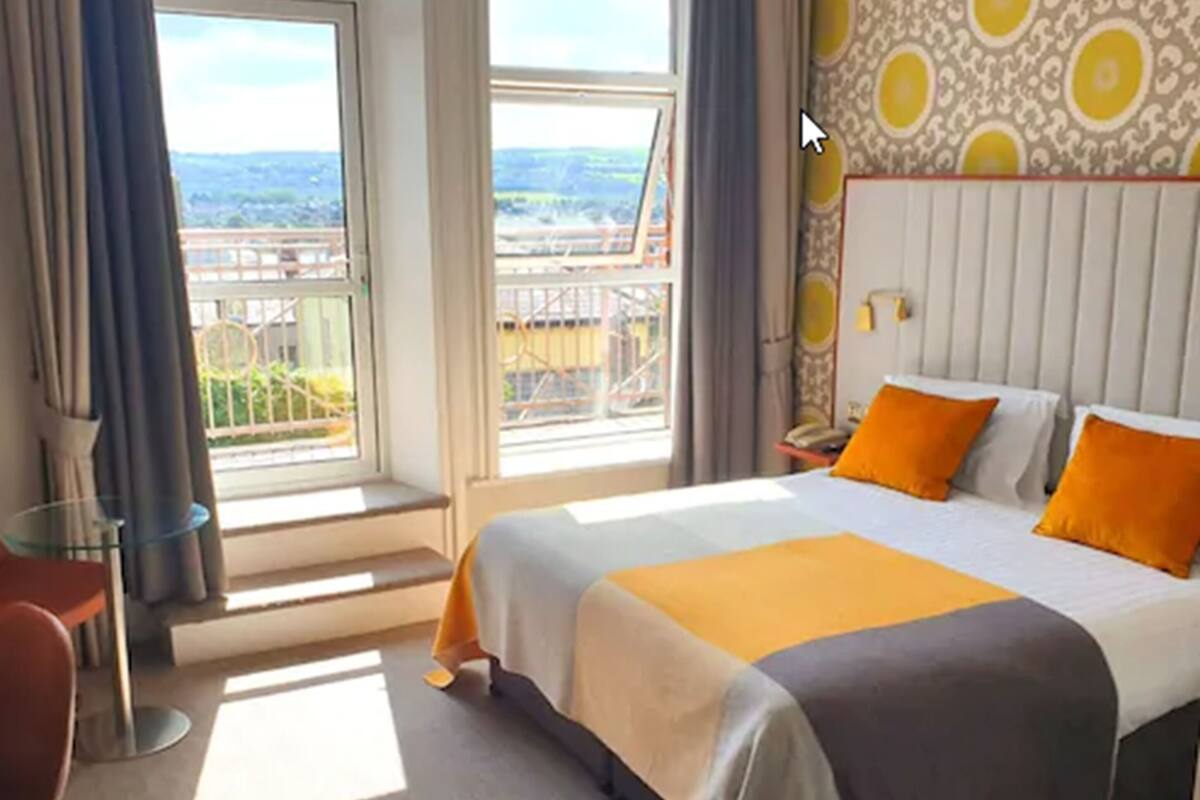 A hotel room with a large bed, two orange pillows, a nightstand with a lamp, a view of a cityscape through a large window, and a patterned wallpaper.