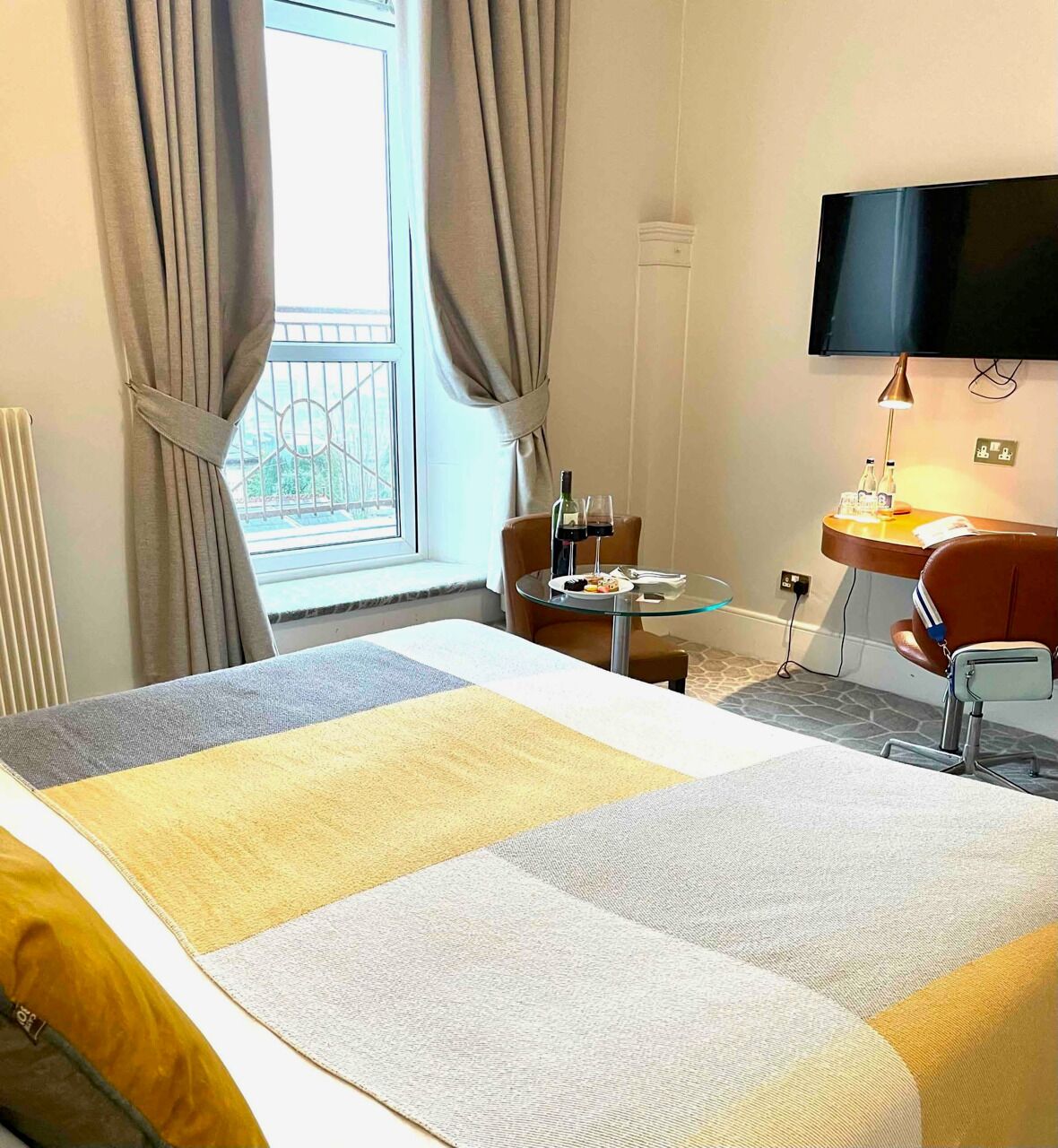 A hotel room with a bed, a small table with a bottle and glass, a chair, a desk with a lamp, and a TV mounted on the wall.