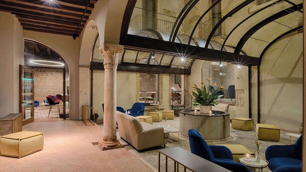 A spacious lobby with a high ceiling, a central seating area with sofas and chairs, a stone wall, and a staircase leading to an upper level. - NH Collection Palazzo Verona (Verona)