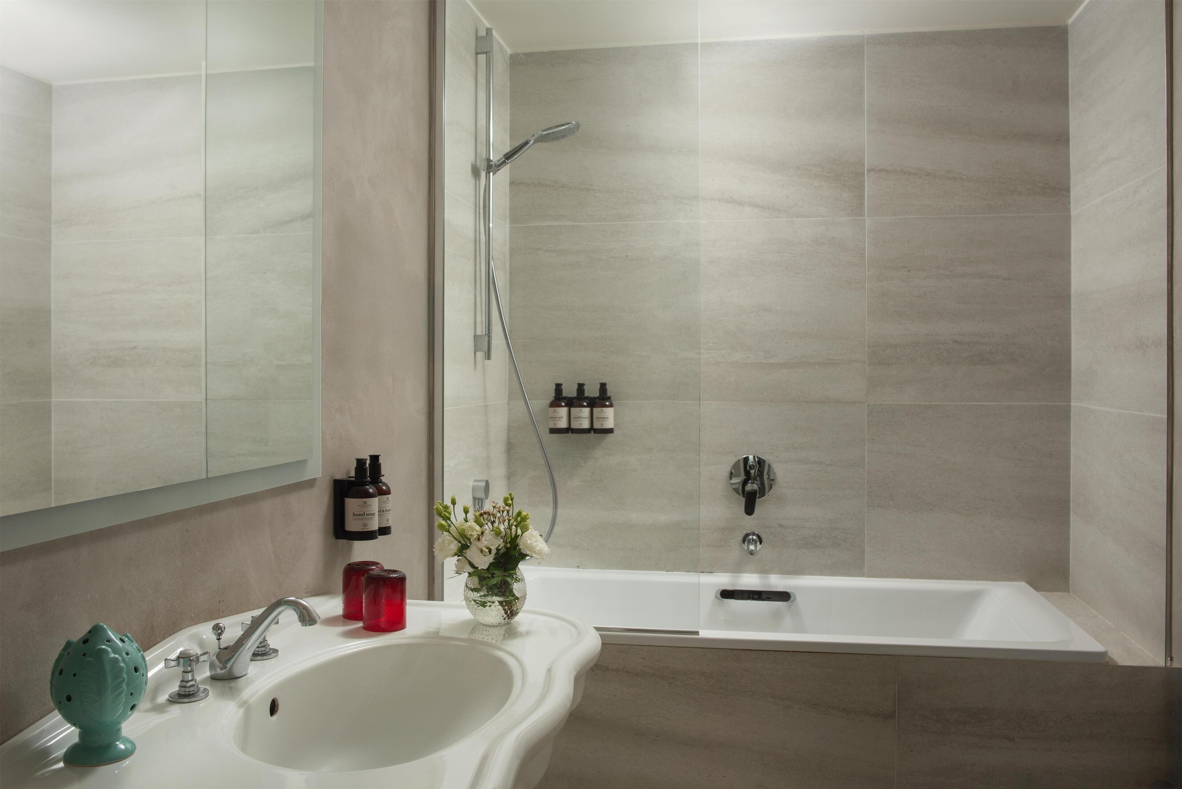 A bathroom with a sink, mirror, shower, and a vase of flowers.