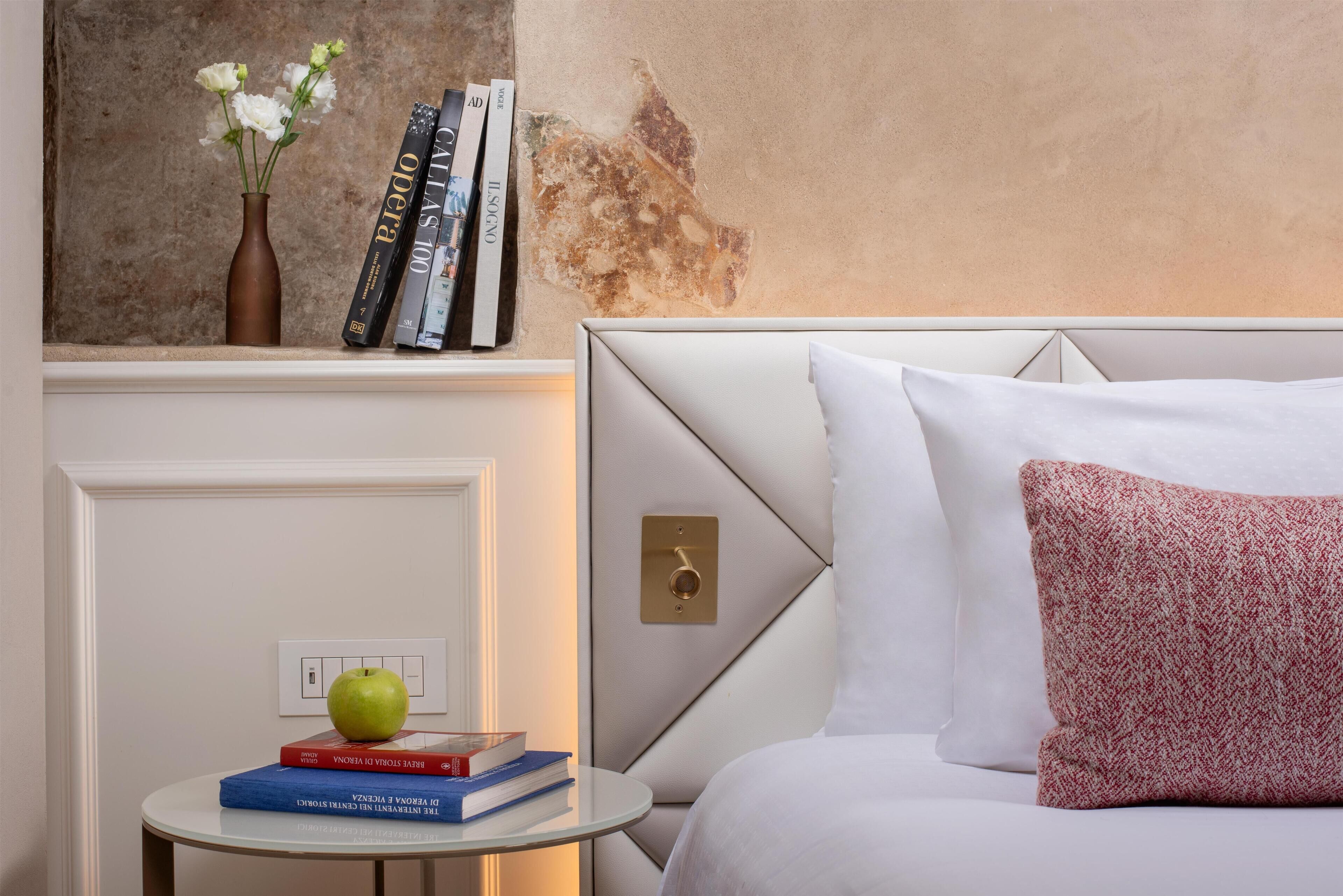 Une chambre avec un lit blanc, une table d’appoint ronde sur laquelle se trouvent des livres et une pomme, un vase rempli de fleurs et un mur dont la peinture s’écaille.