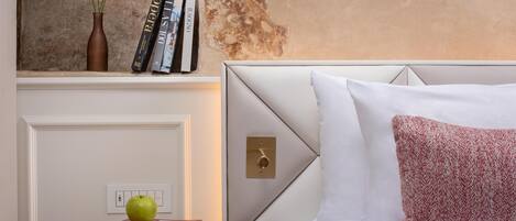 Un dormitorio con una cama blanca, una mesita redonda con libros y una manzana, un jarrón con flores, y una pared con pintura descascarada.