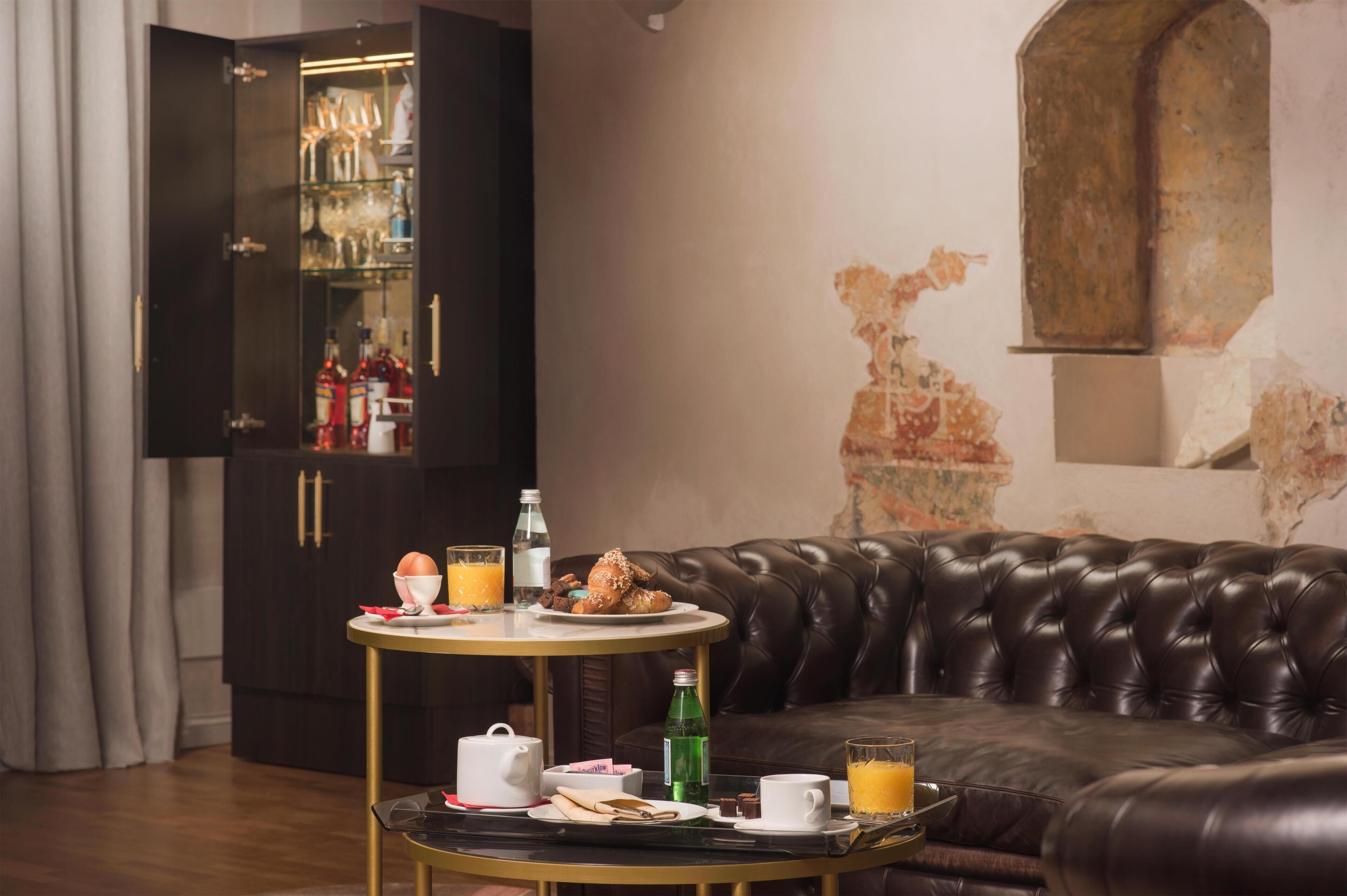 A room with a leather sofa, a round table with breakfast items, a cabinet with bottles, and a wall with a decorative arch.