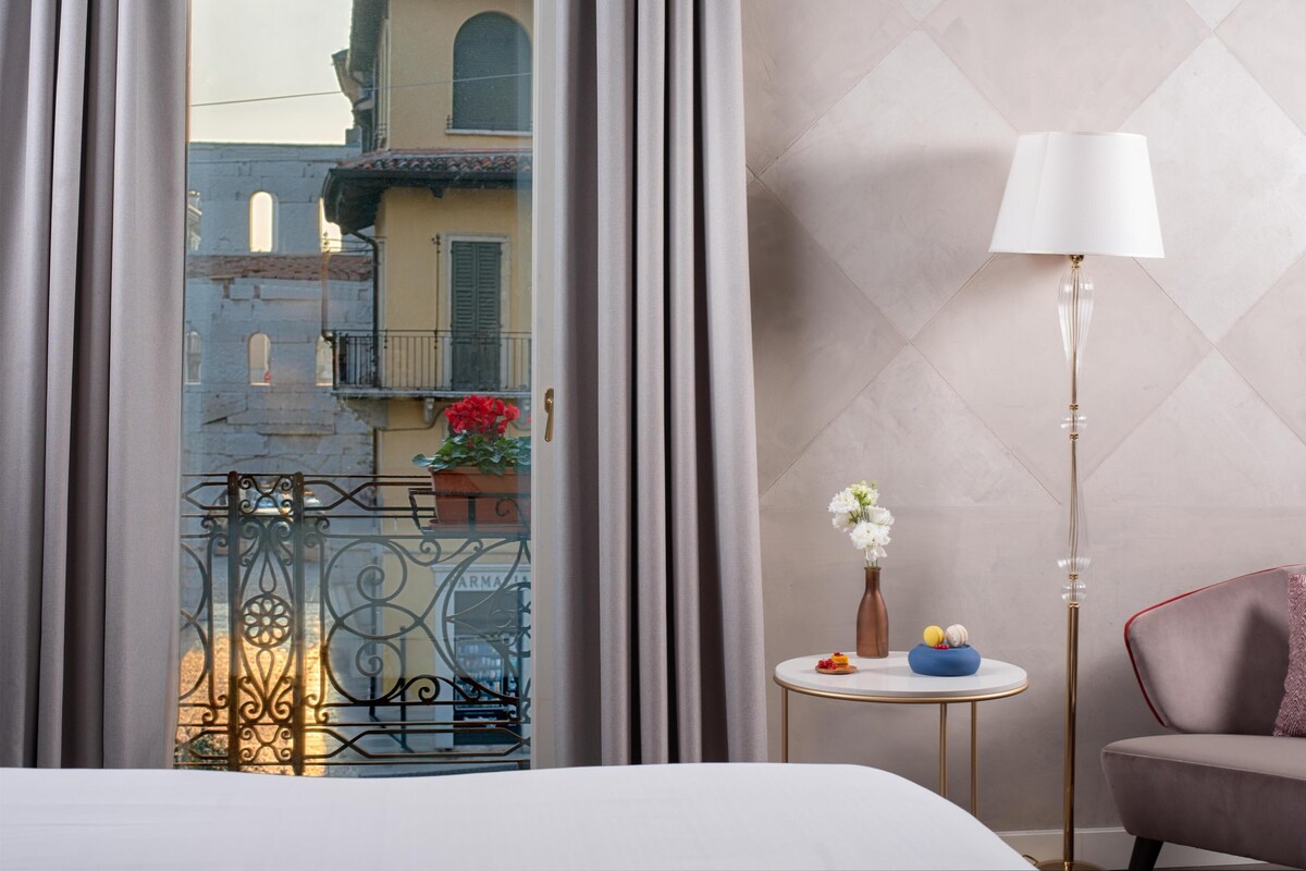 Une chambre d’hôtel avec vue sur un balcon, une table ronde ornée d’un vase et de fruits, un lampadaire et un lit.