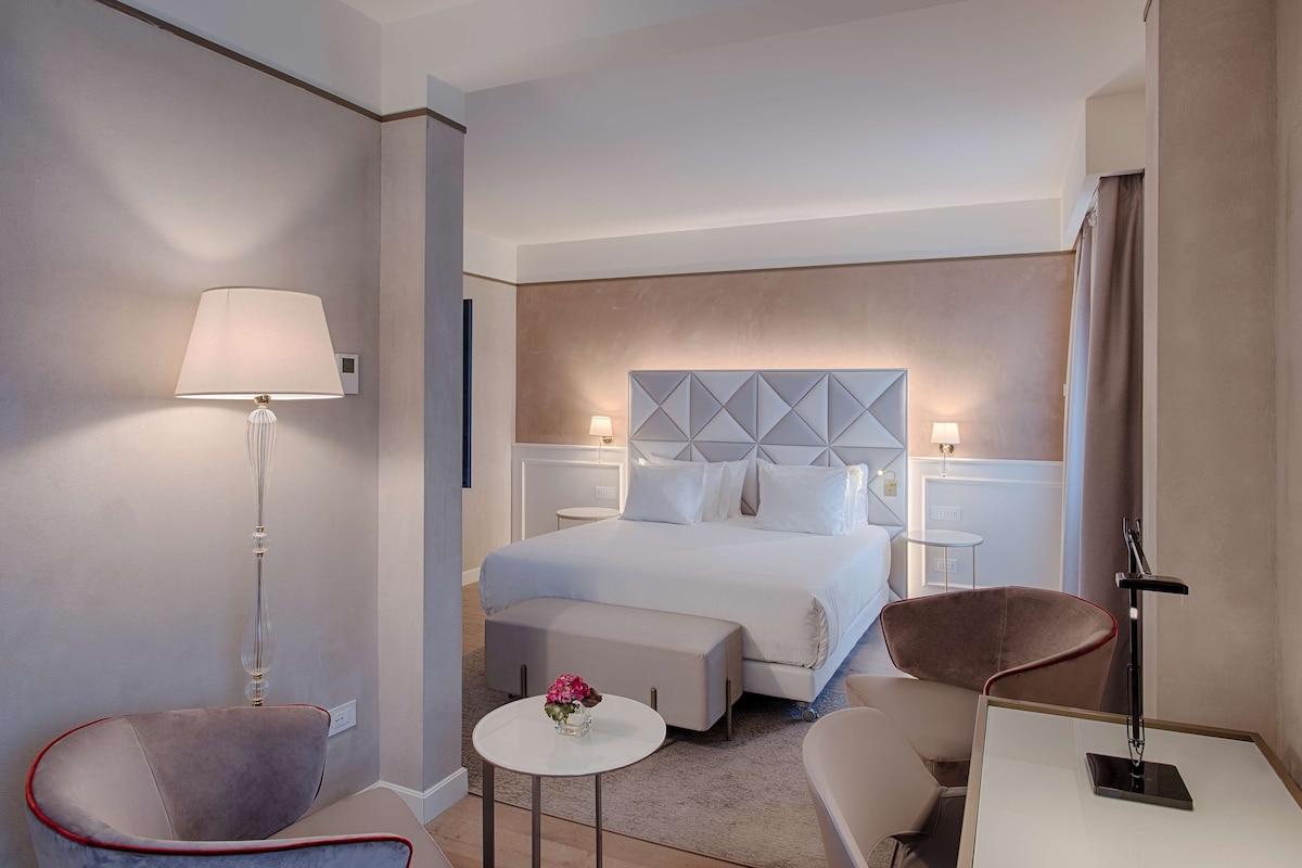 A hotel room with a bed, two chairs, a small table, and a lamp.