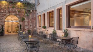 An outdoor café with stone walls, metal tables, and chairs, featuring a brick archway and large windows. - NH Collection Palazzo Verona (Verona)