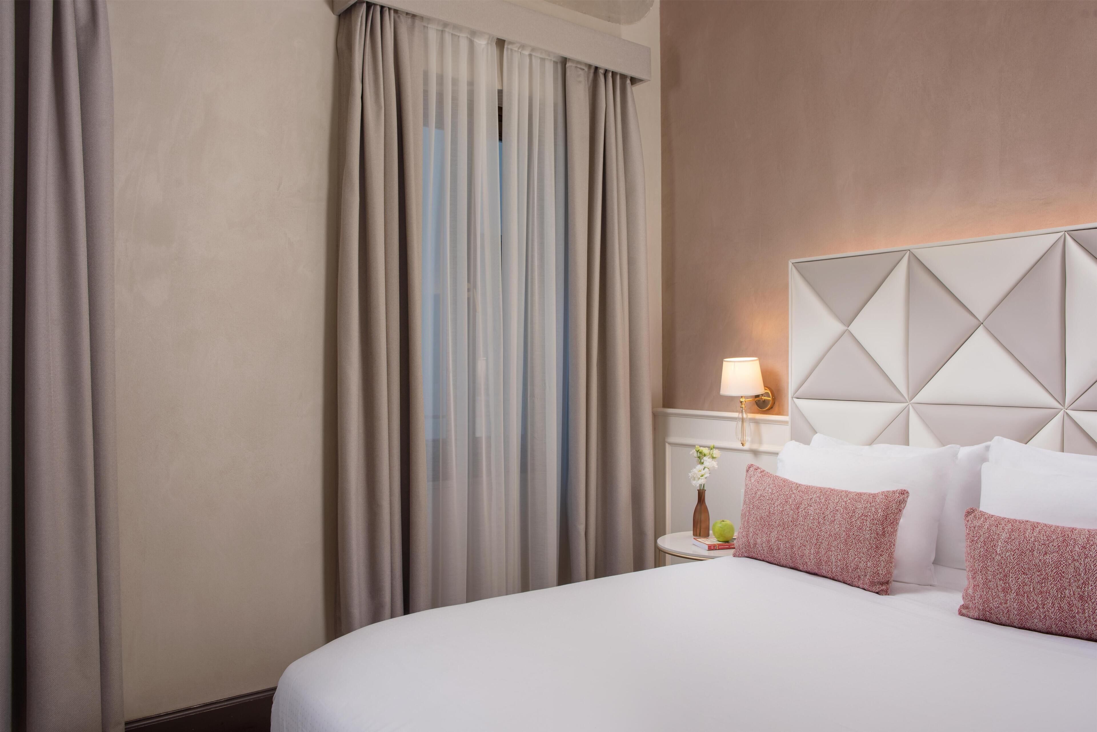 A hotel room with a bed, curtains, a bedside table with a lamp, and a decorative wall panel.