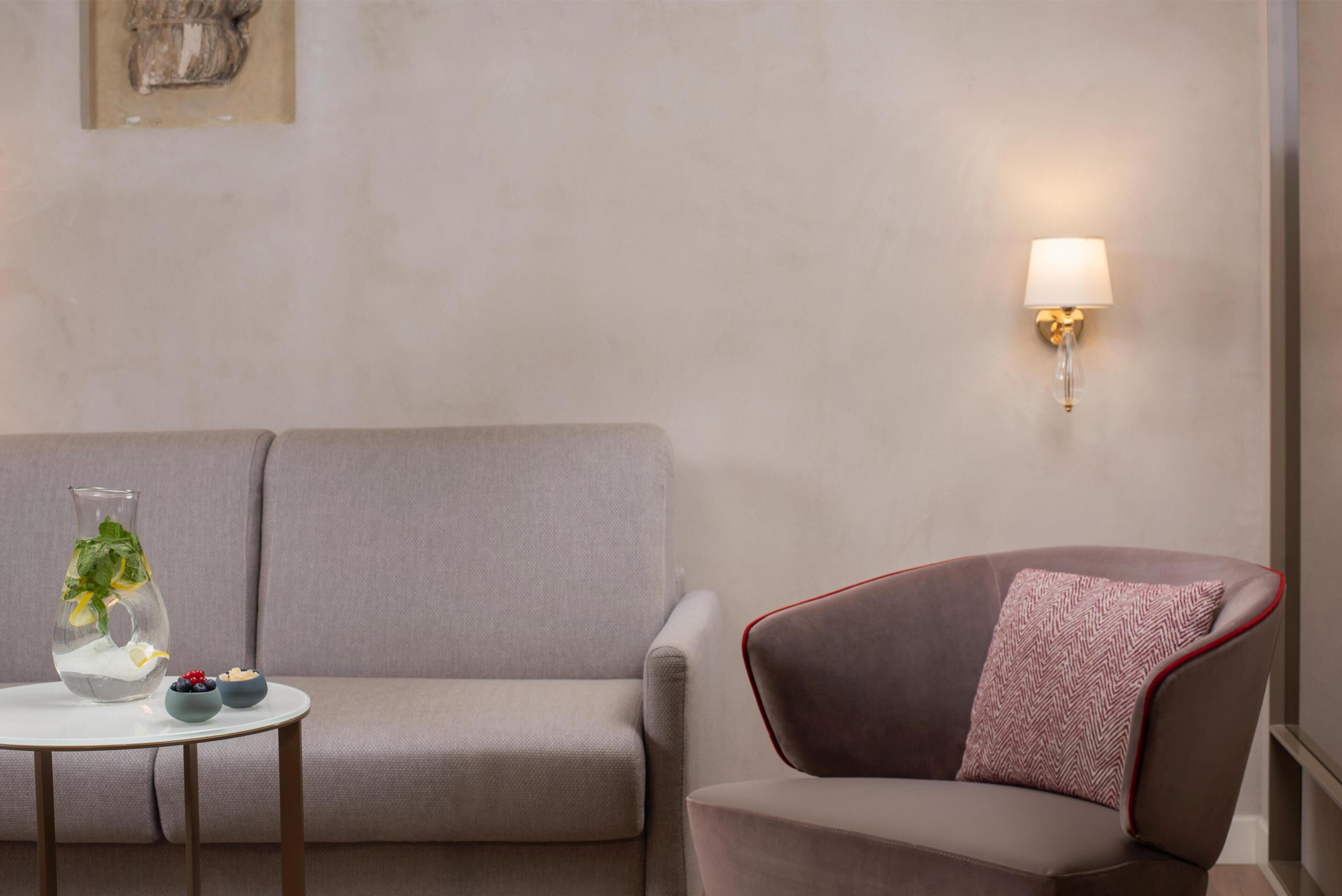 A neutral-toned room with a sofa, a small round table with a vase and bowls, and a wall-mounted lamp.