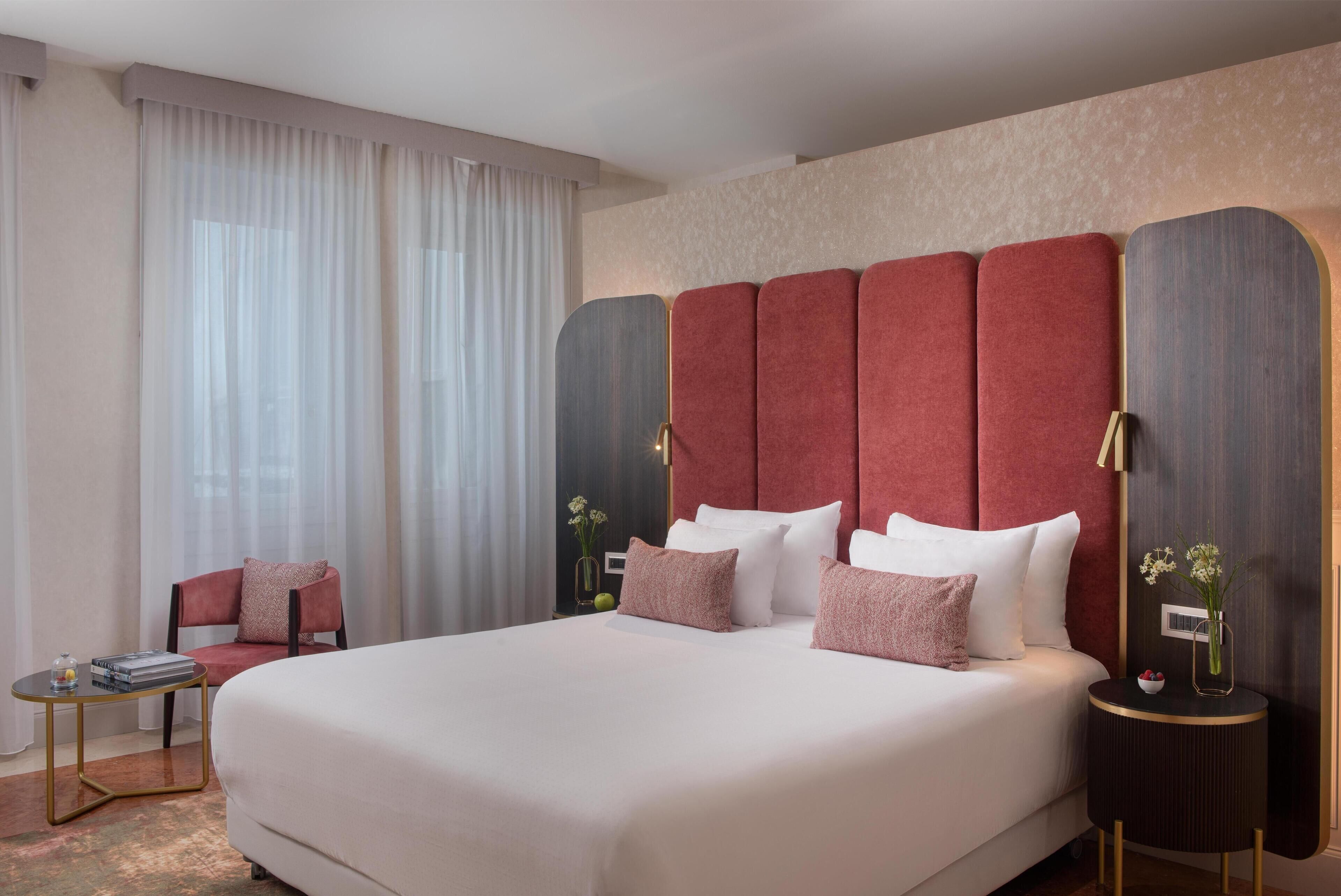 Camera d'albergo con un letto grande, una poltrona imbottita rossa, un tavolino rotondo e un comodino con un vaso di fiori.