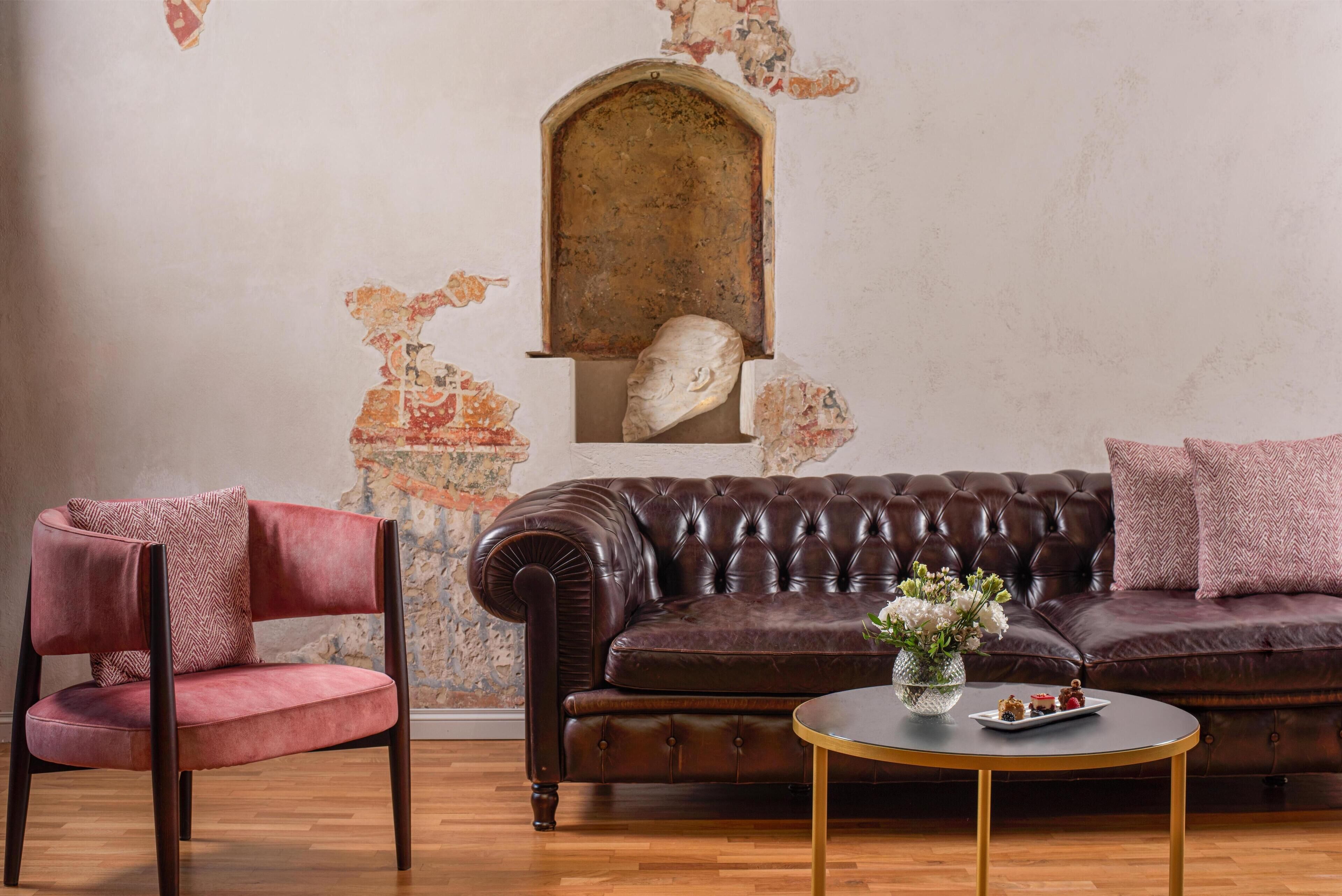 A room with a brown leather sofa, a red velvet armchair, a small round table with a vase, and a wall with a decorative archway.