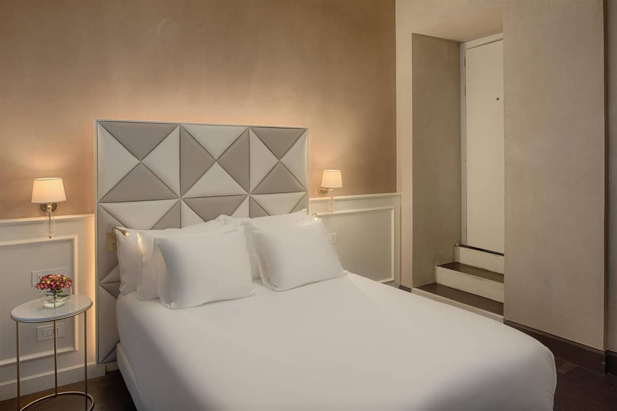 Une chambre d’hôtel avec un lit, deux lampes de chevet, une petite table sur laquelle est posé un vase de fleurs, et une porte qui donne sur une autre pièce.