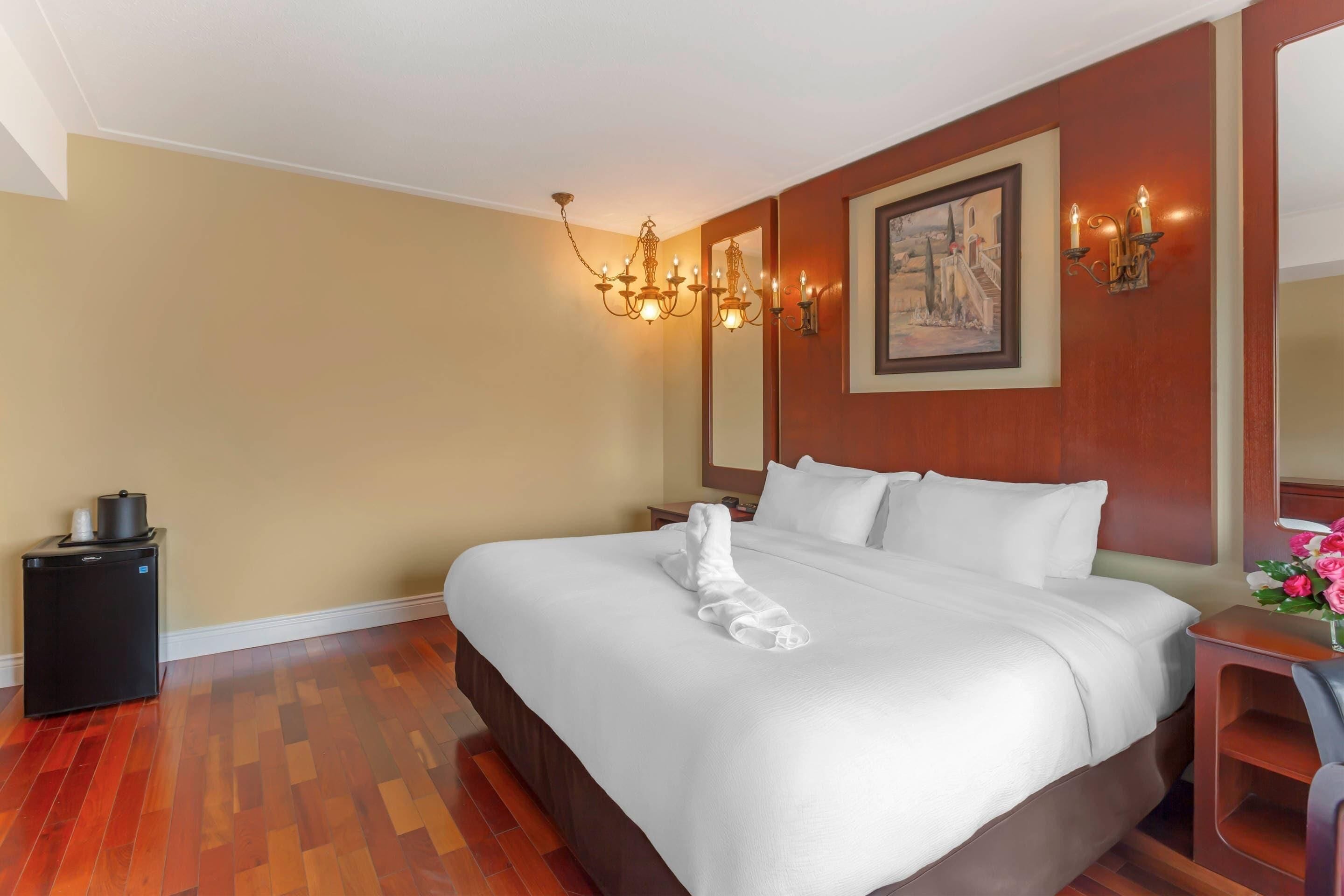 A hotel room with a large bed, a bedside table, a small desk, a chair, a mirror, a chandelier, and a painting on the wall.