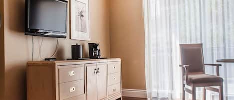 A hotel room with a flat-screen TV mounted on the wall, a wooden cabinet with drawers, a coffee maker, and a chair near a window with sheer curtains.