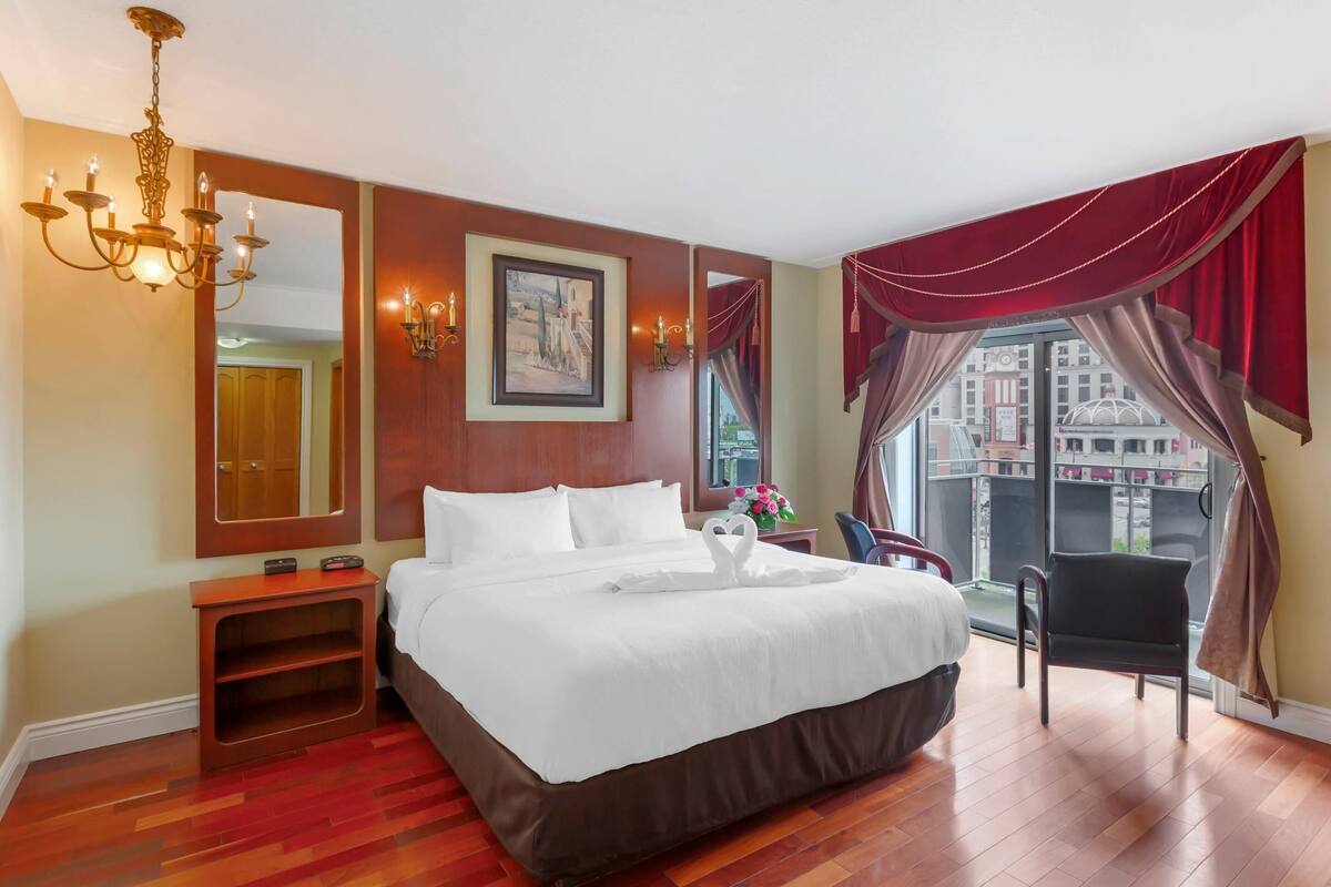 A hotel room with a large bed, a wooden nightstand, a mirror, a chandelier, a window with curtains, and a view of buildings outside.