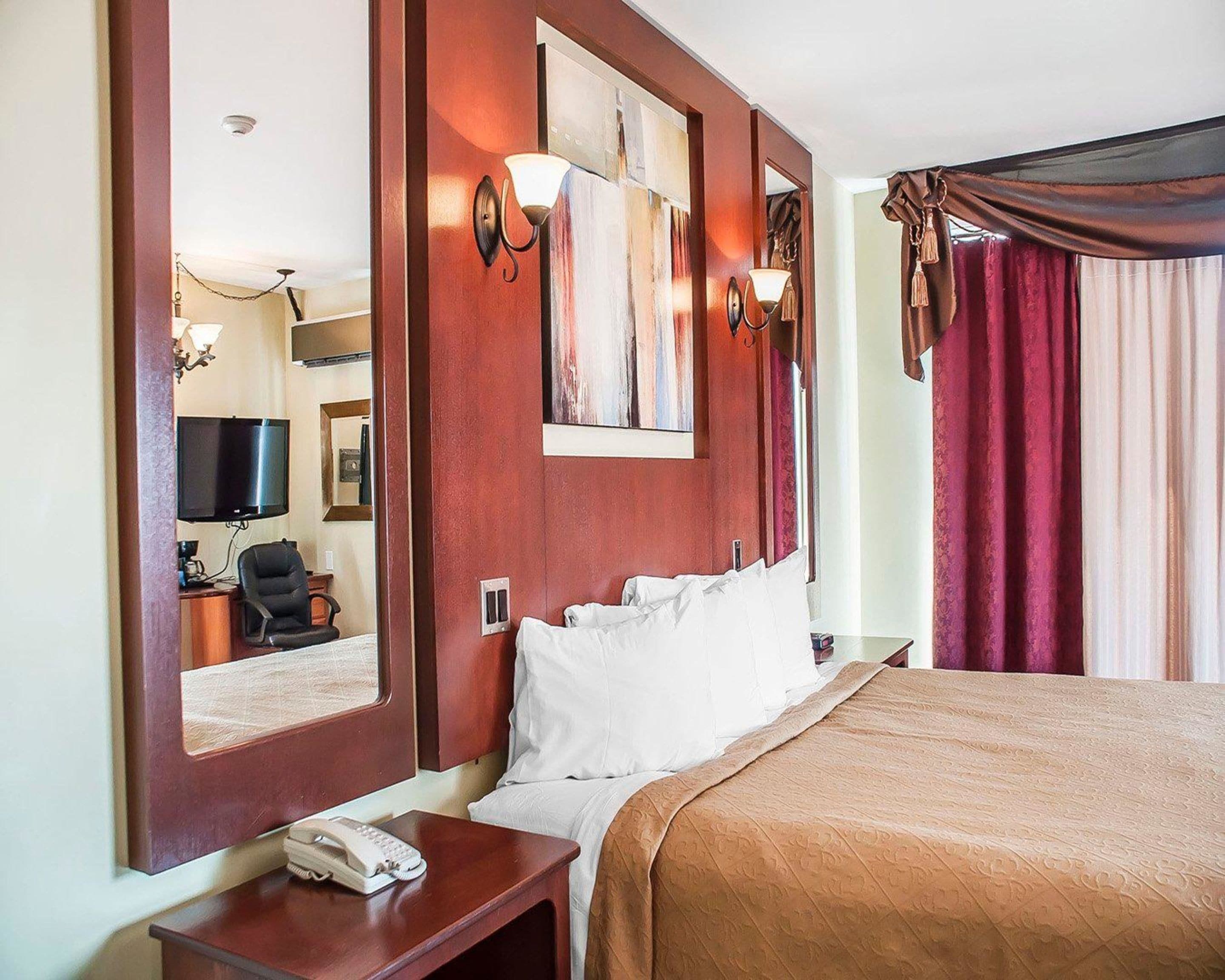A hotel room with a large bed, a bedside table with a telephone, a mirror, and a television on the wall.