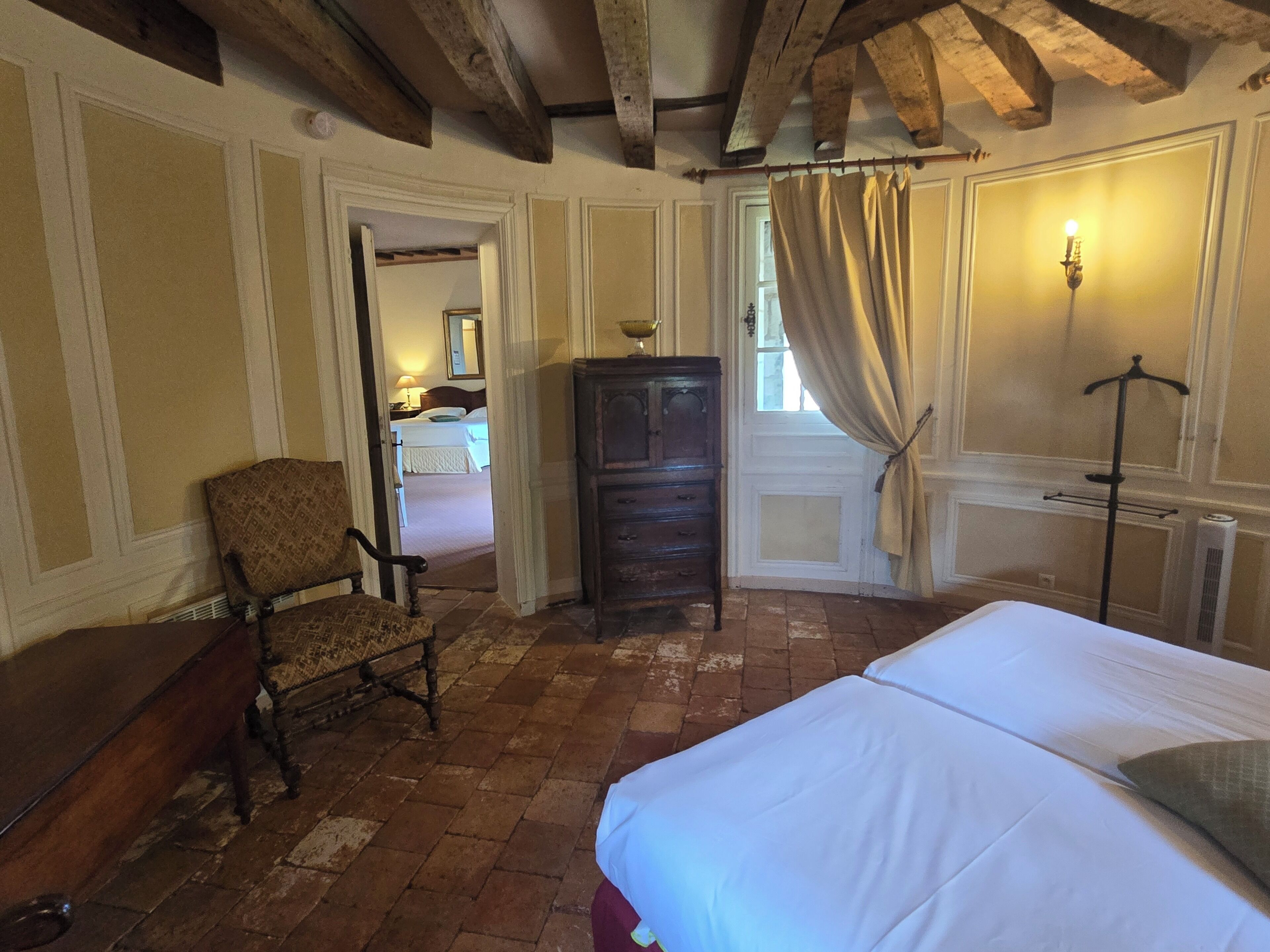 Un dormitorio con una cama grande, suelo de madera, una silla, un aparador y una ventana con cortinas.